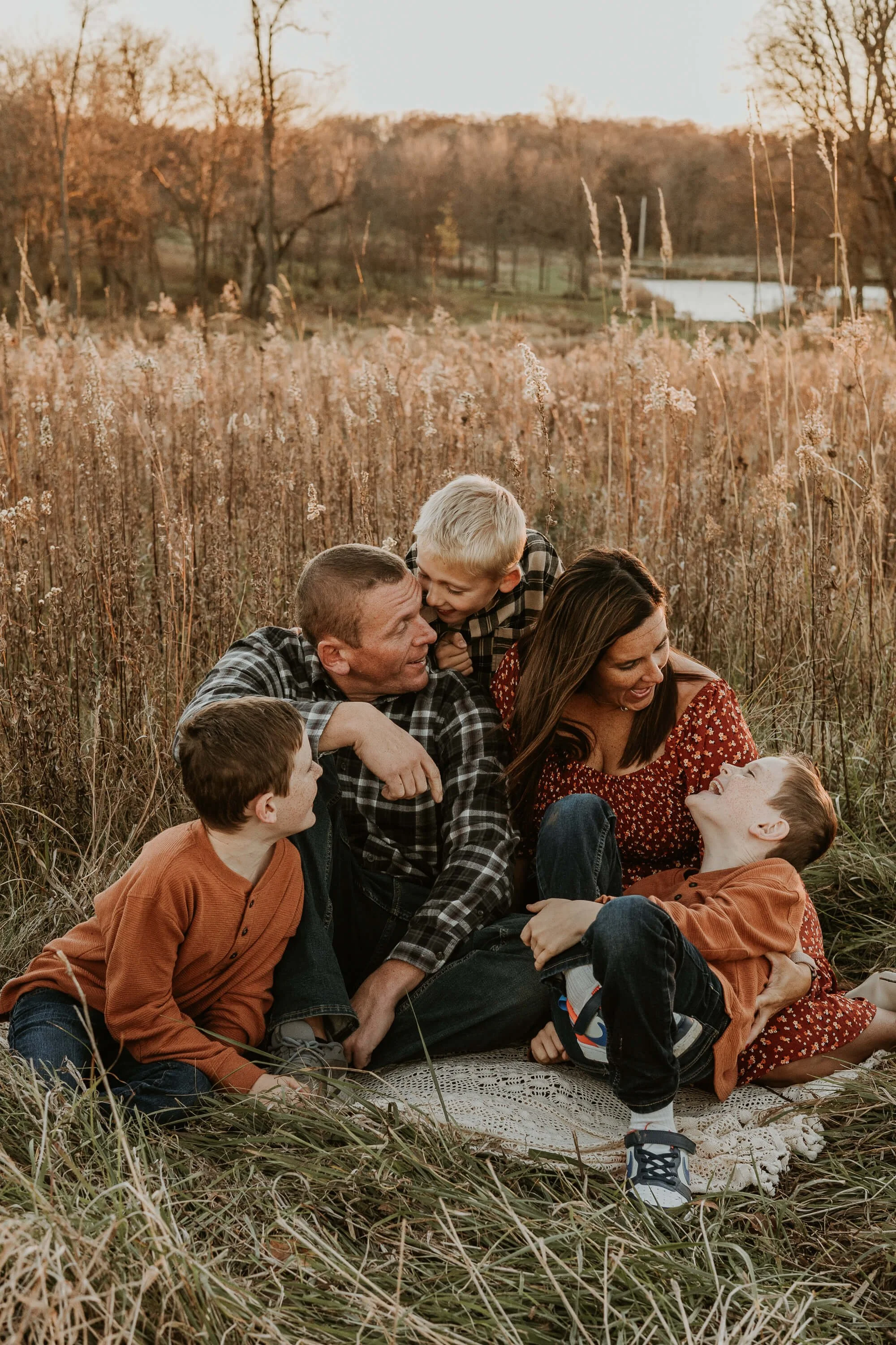 A family of five - dad, mom, and three brothers - laugh playfully on a blanket in a field of prairie grass