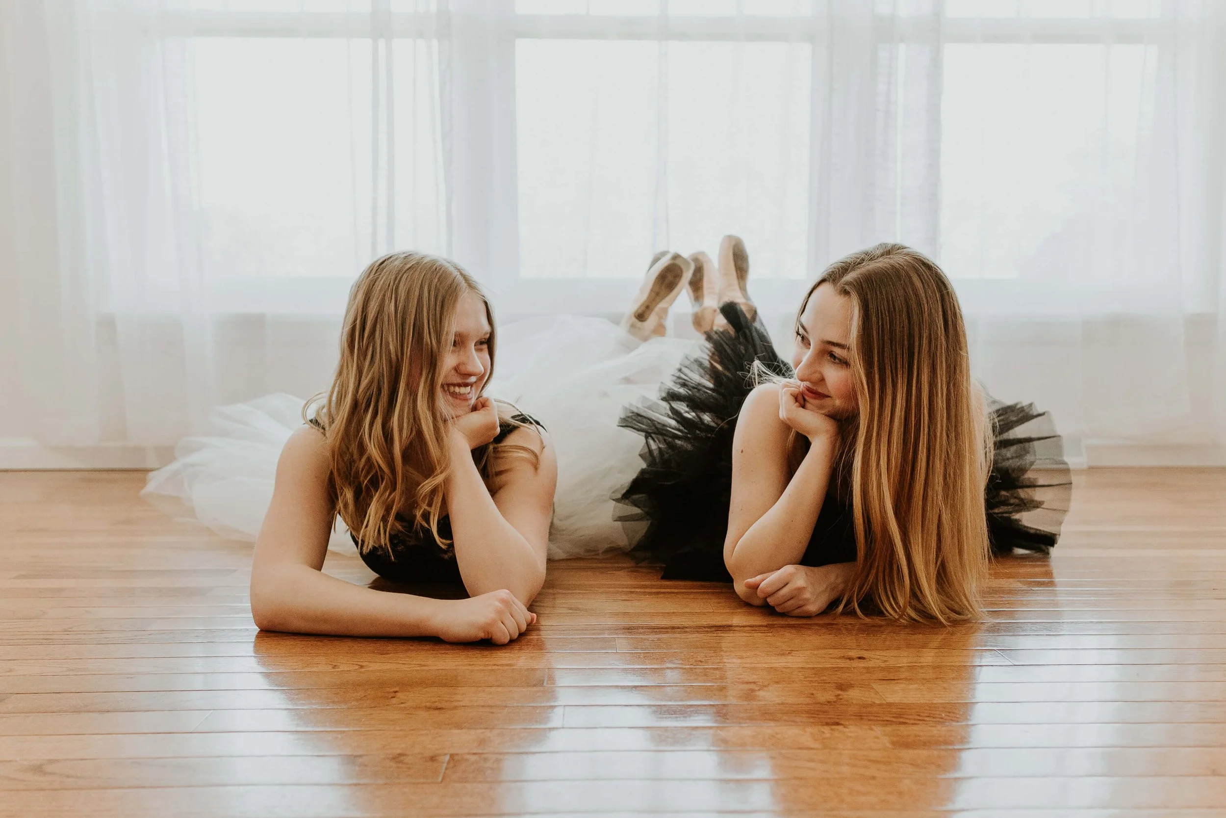 Two friends, both dancers, lay on the floor with their hands supporting their chins, knowing smiles on their faces as they look at one another.
