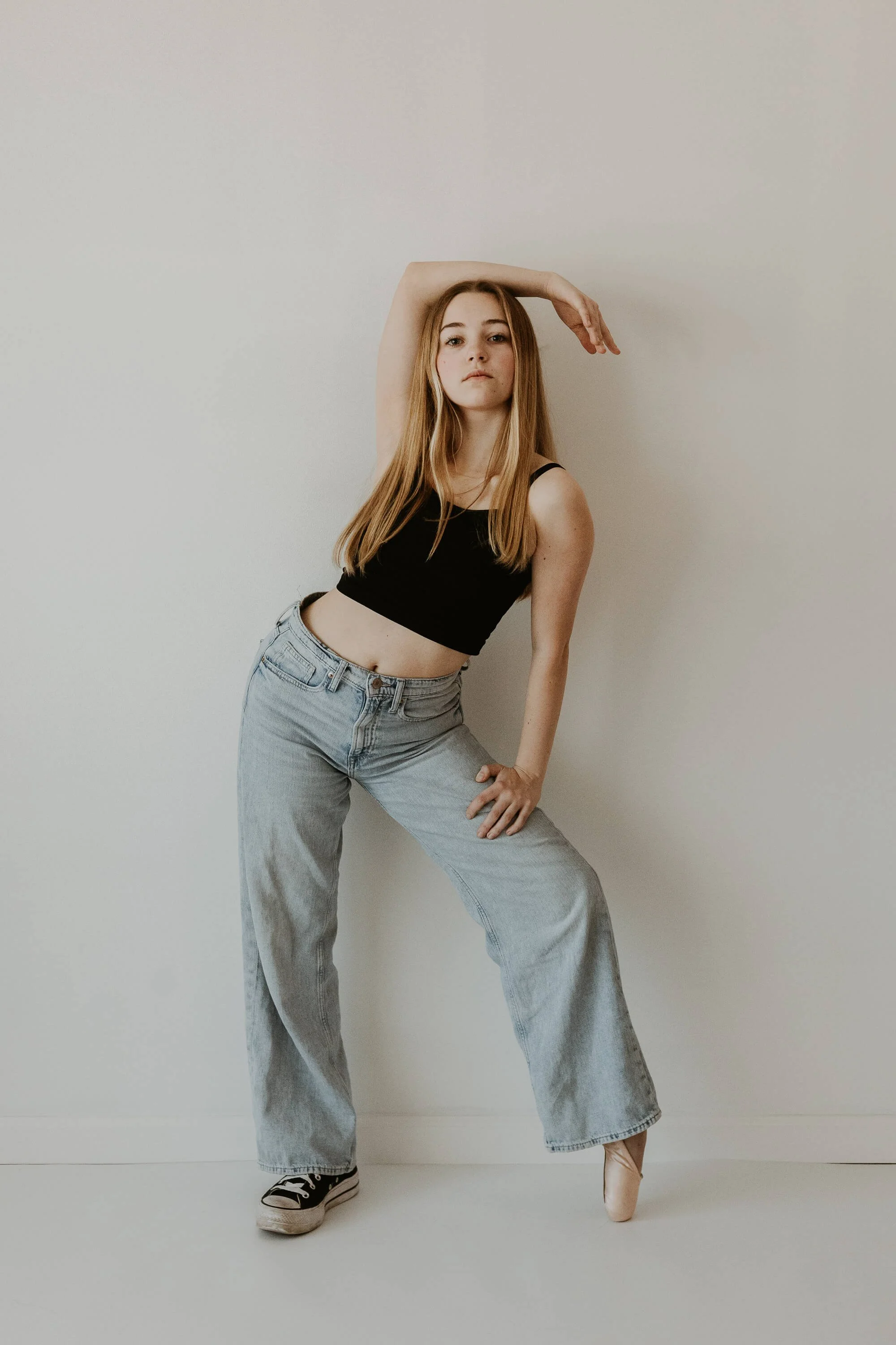 A dancer holding an exaggerated pose, a hip jutting out to the left, one arm resting atop her hed, the other hand placed on her leg, which is up en pointe.