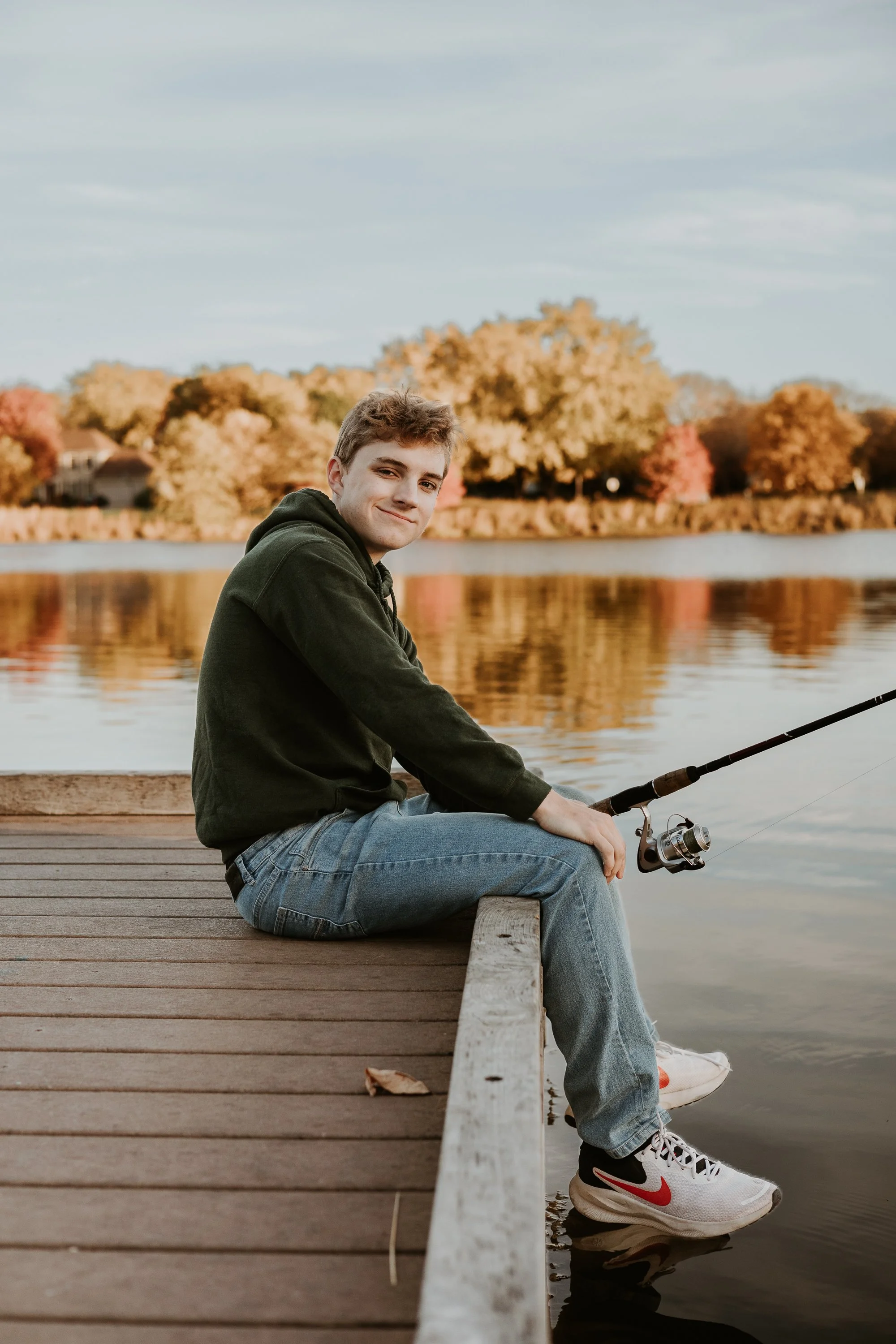A senior boy from Tri Valley High School sits at the end of a dock by a lake, with a fishing pole in hand.