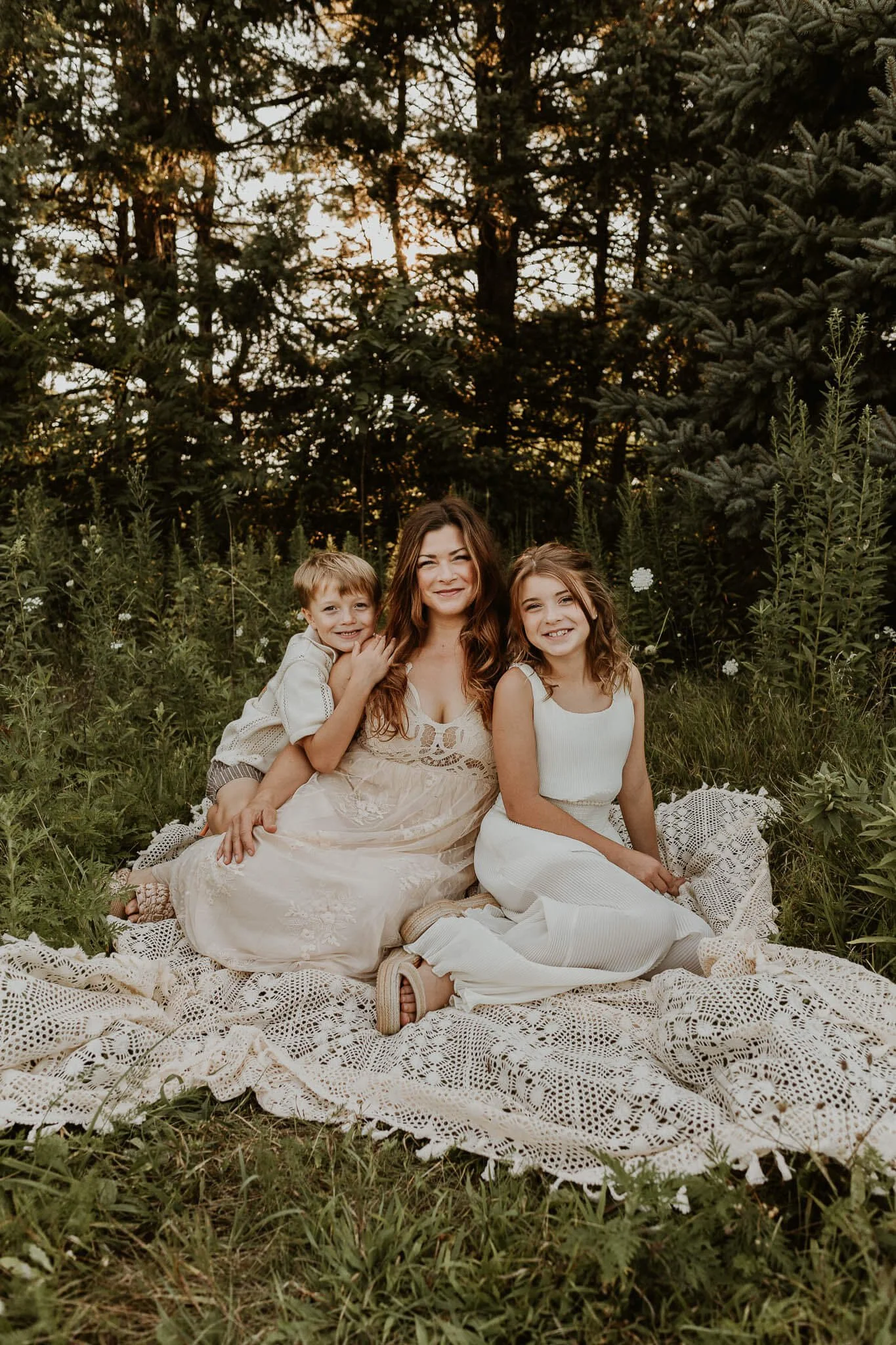 A mother and her 2 children sit on a blanket smiling as they cuddle together