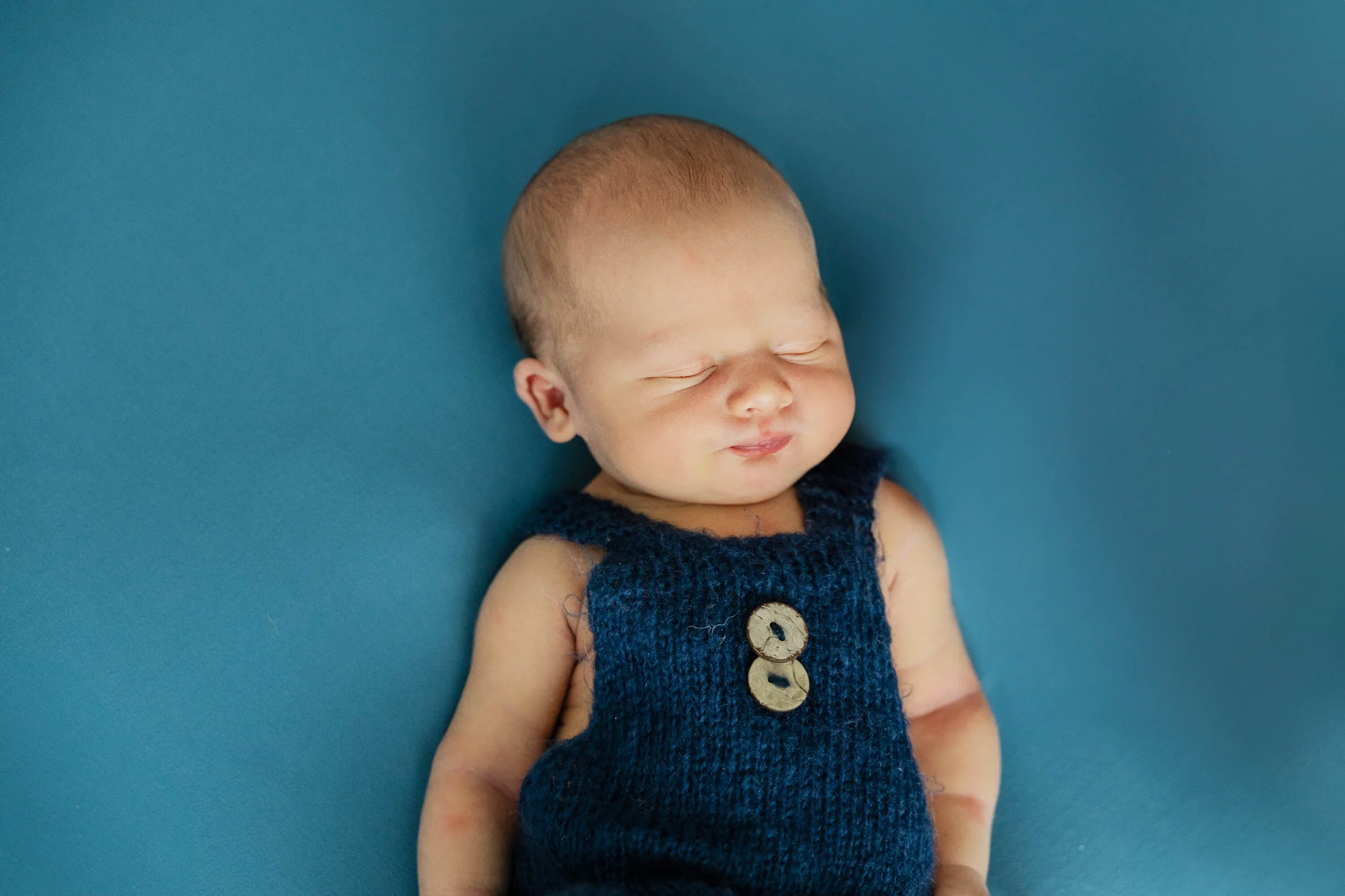 A baby sleeps peacefully wearing knitted overalls against a blue backdrop