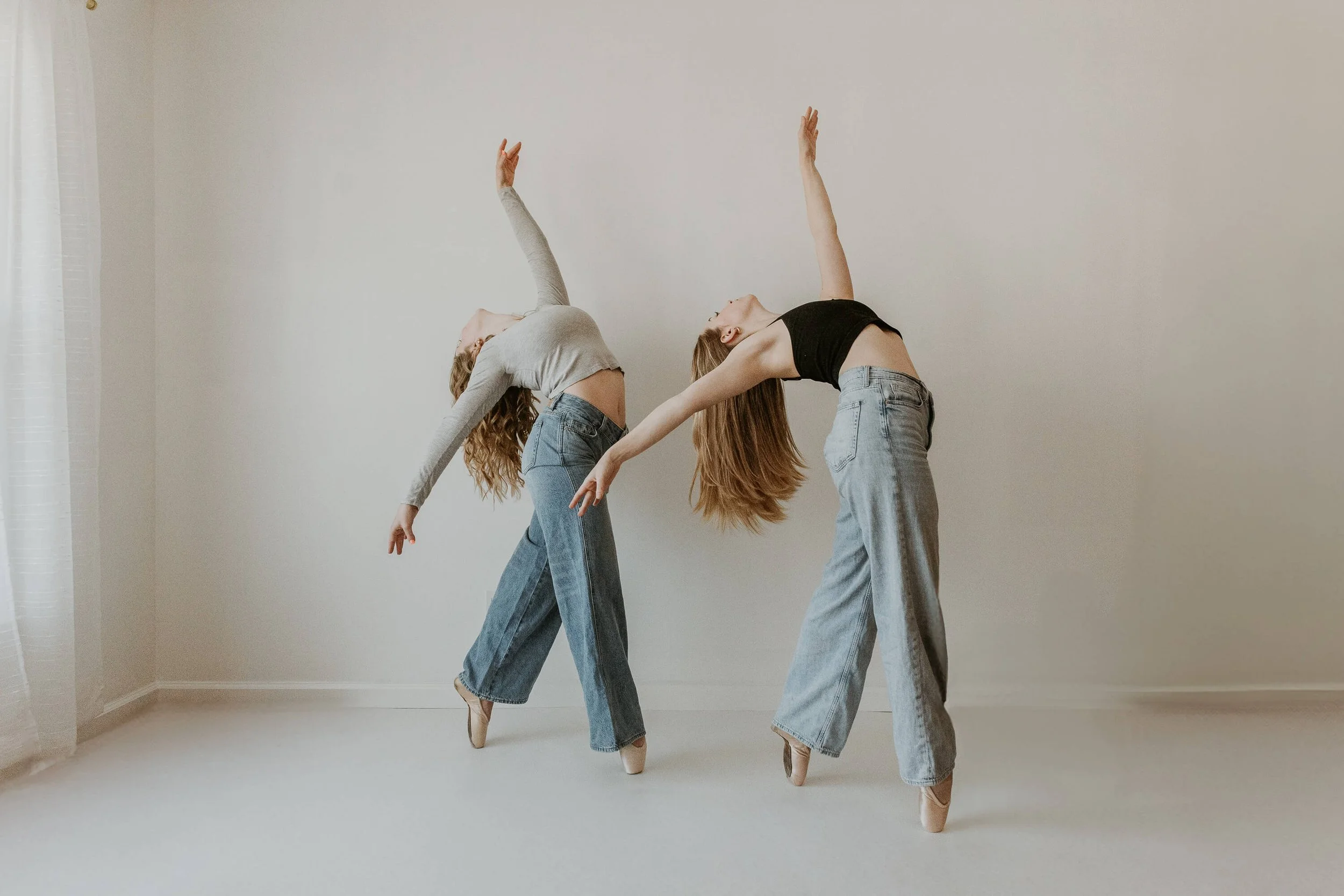 Two ballet dancers hold the same pose en pointe, one arm stretched above and the other dropped gracefully behind, as they arch their backs.