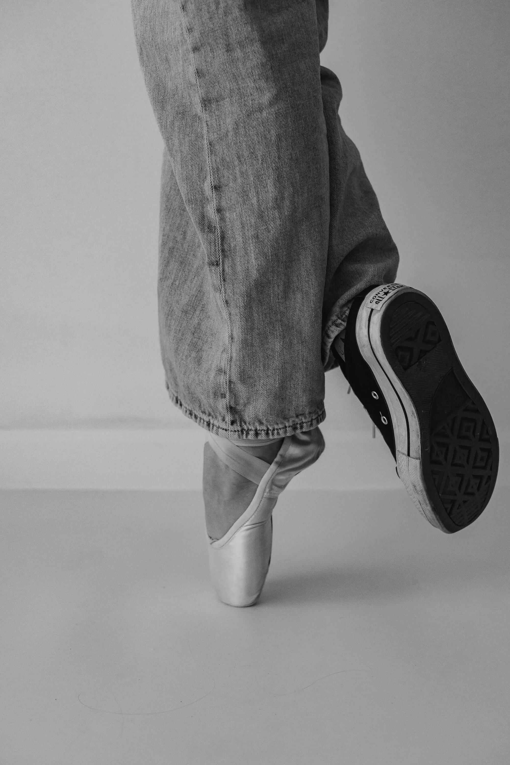 Up close, black and white of a dancer wearing one sneaker and one pointe shoe.