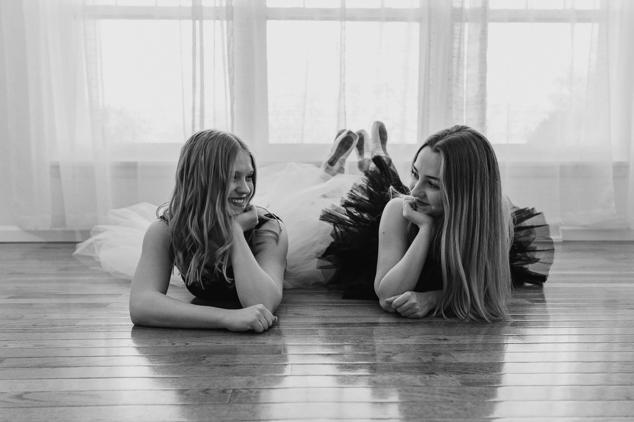 Two high school dancers smile at each other as they lay on their stomachs on the floor.