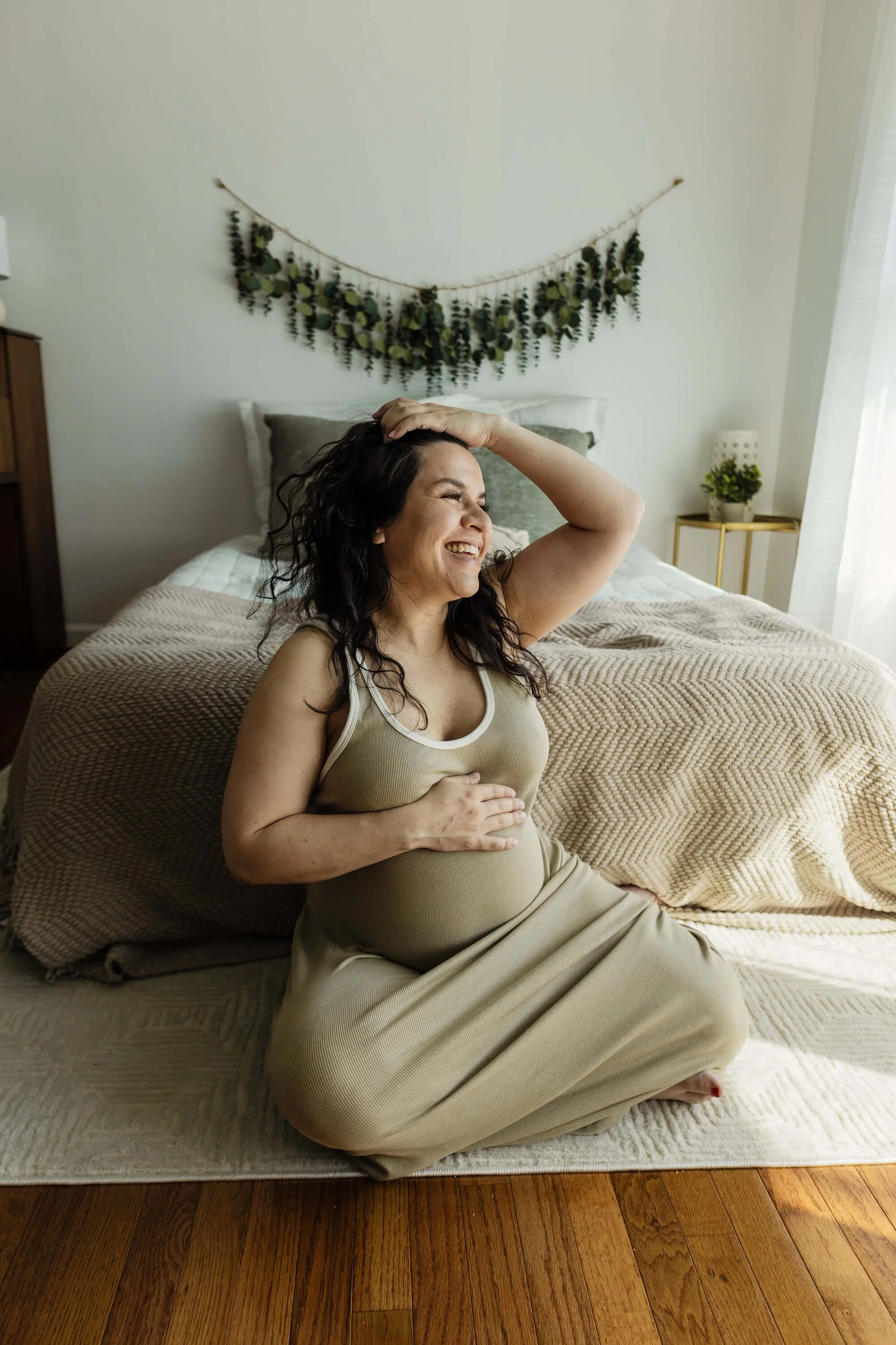 A woman laughs while tossing her hair at the foot of a bed, hand placed gently atop her pregnant belly