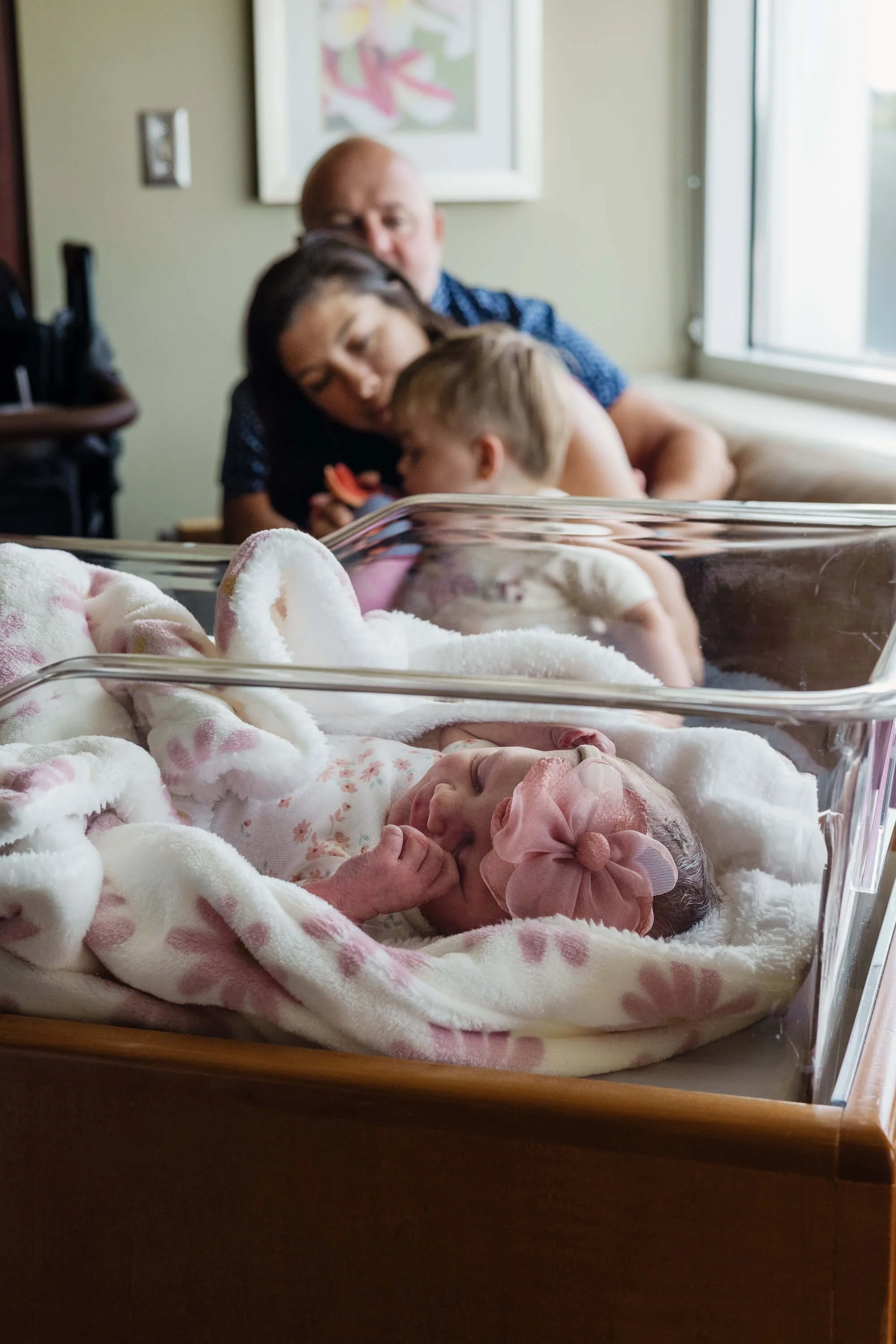 A newborn baby girl can be seen sleeping in a cot while her big brother snuggles on his mother's lap in the background