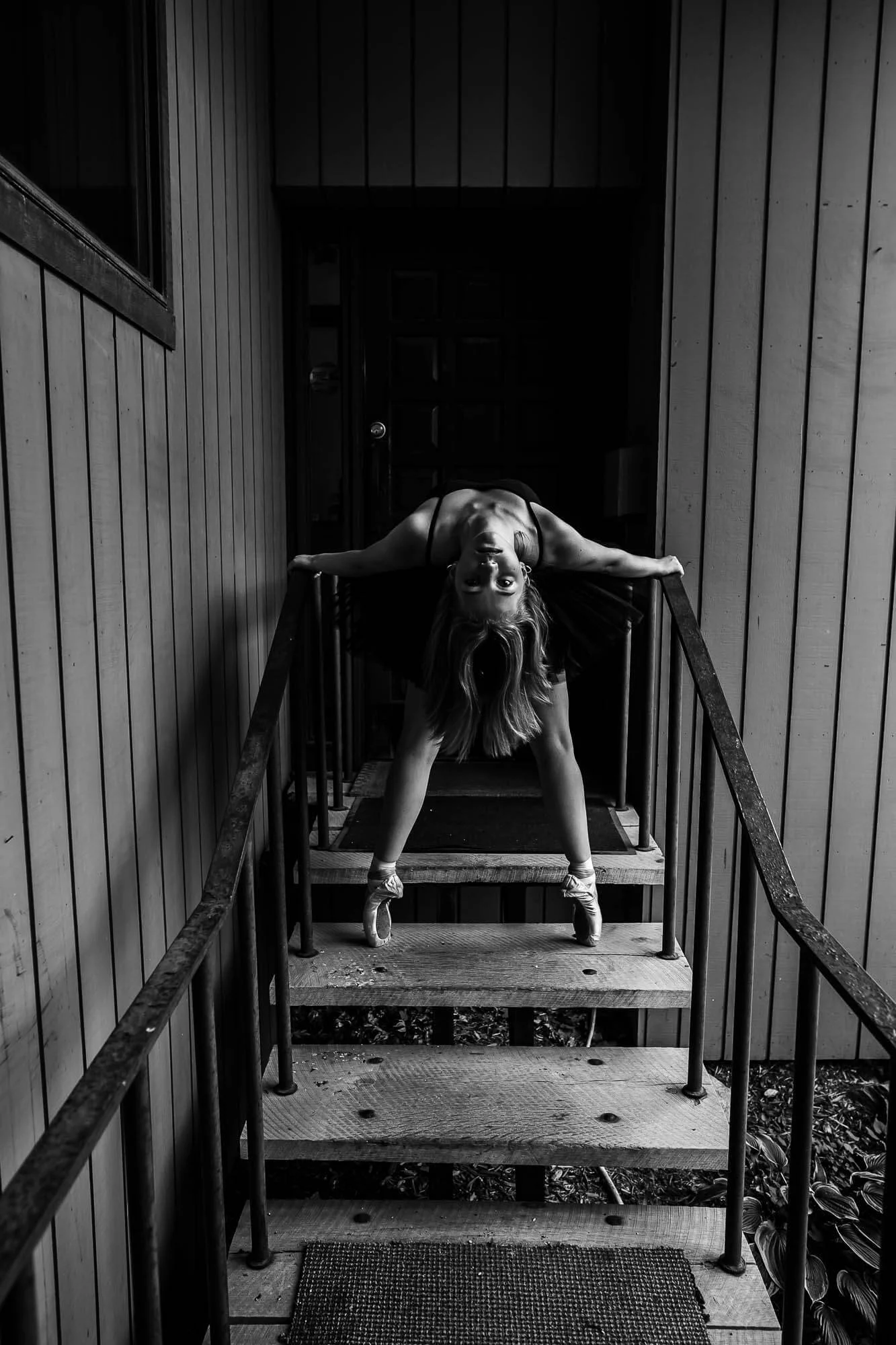 A ballerina stands en pointe while bending back in a fierce pose at a wooden stairway