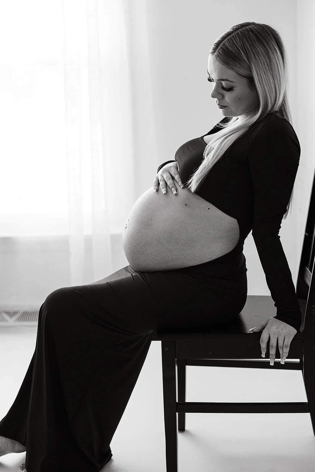 A pregnant mother sits at the edge of a wooden chair, tendlery touching her belly.