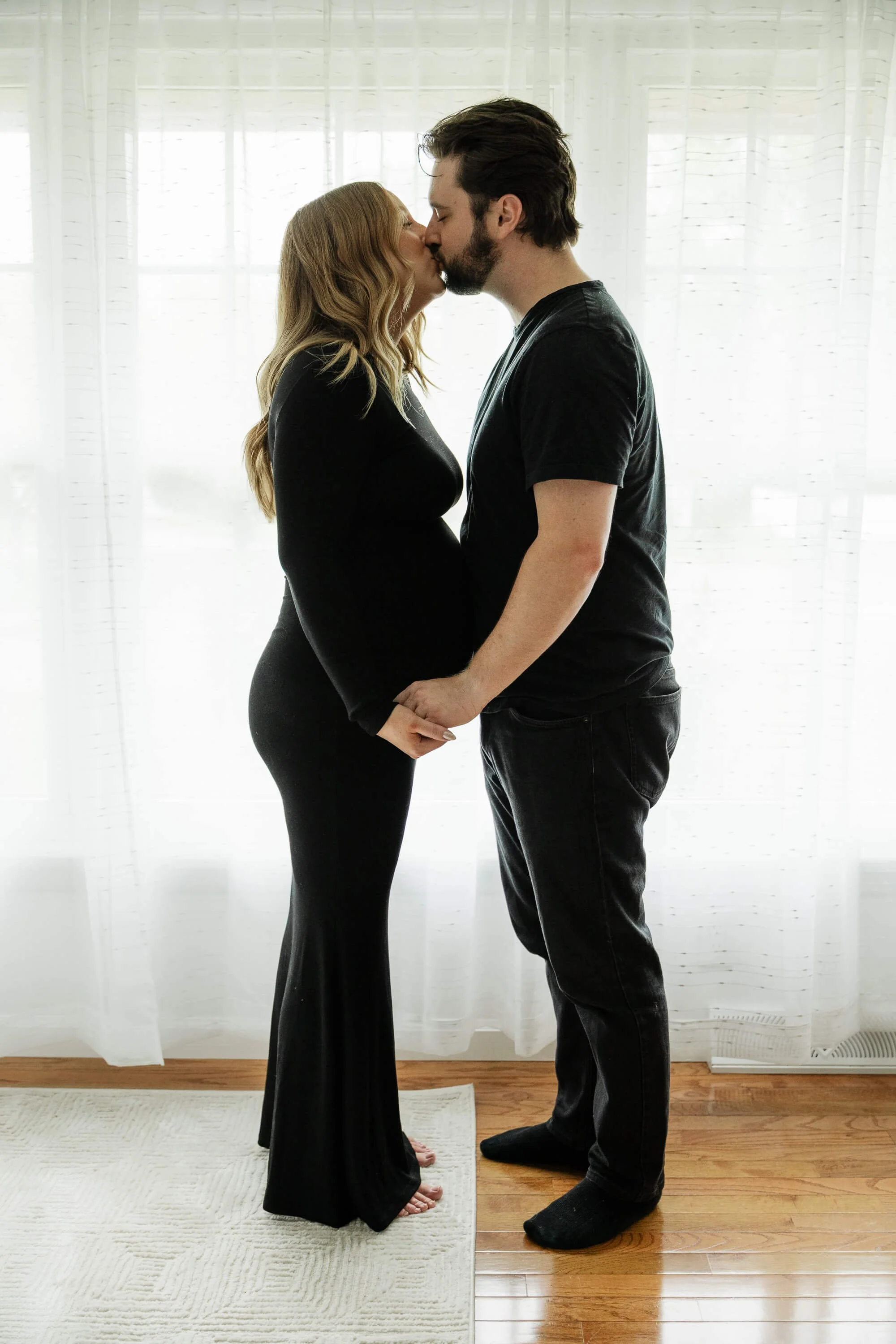 A couple plants a soft kiss on one anothers lips in front of a window during their maternity session at the studio of Liz Walsh Photography