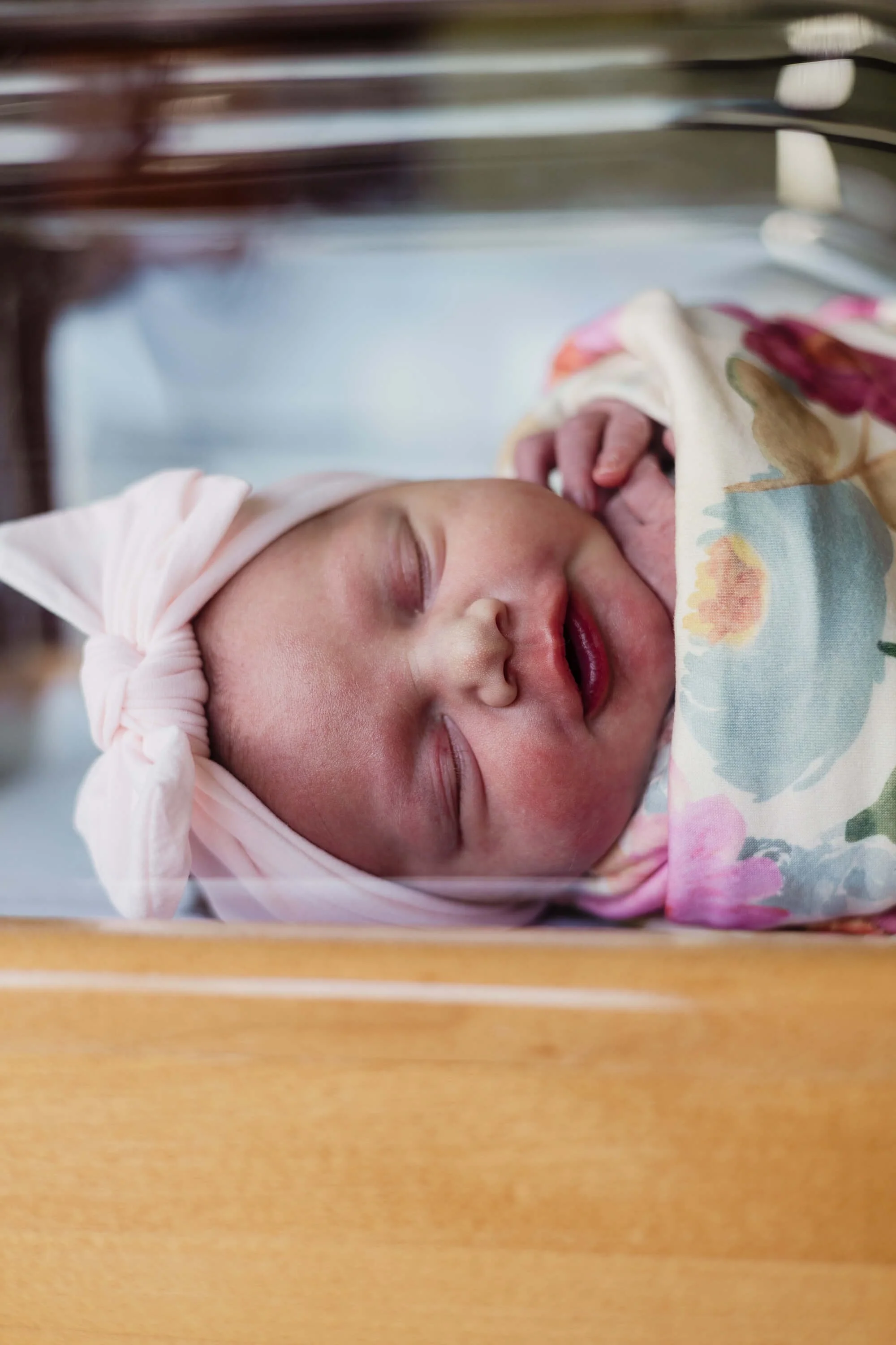A newborn baby sleeps swaddled in a floral wrap with a pink head band in a hospital cot at Carle BroMenn Hospital in Normal IL