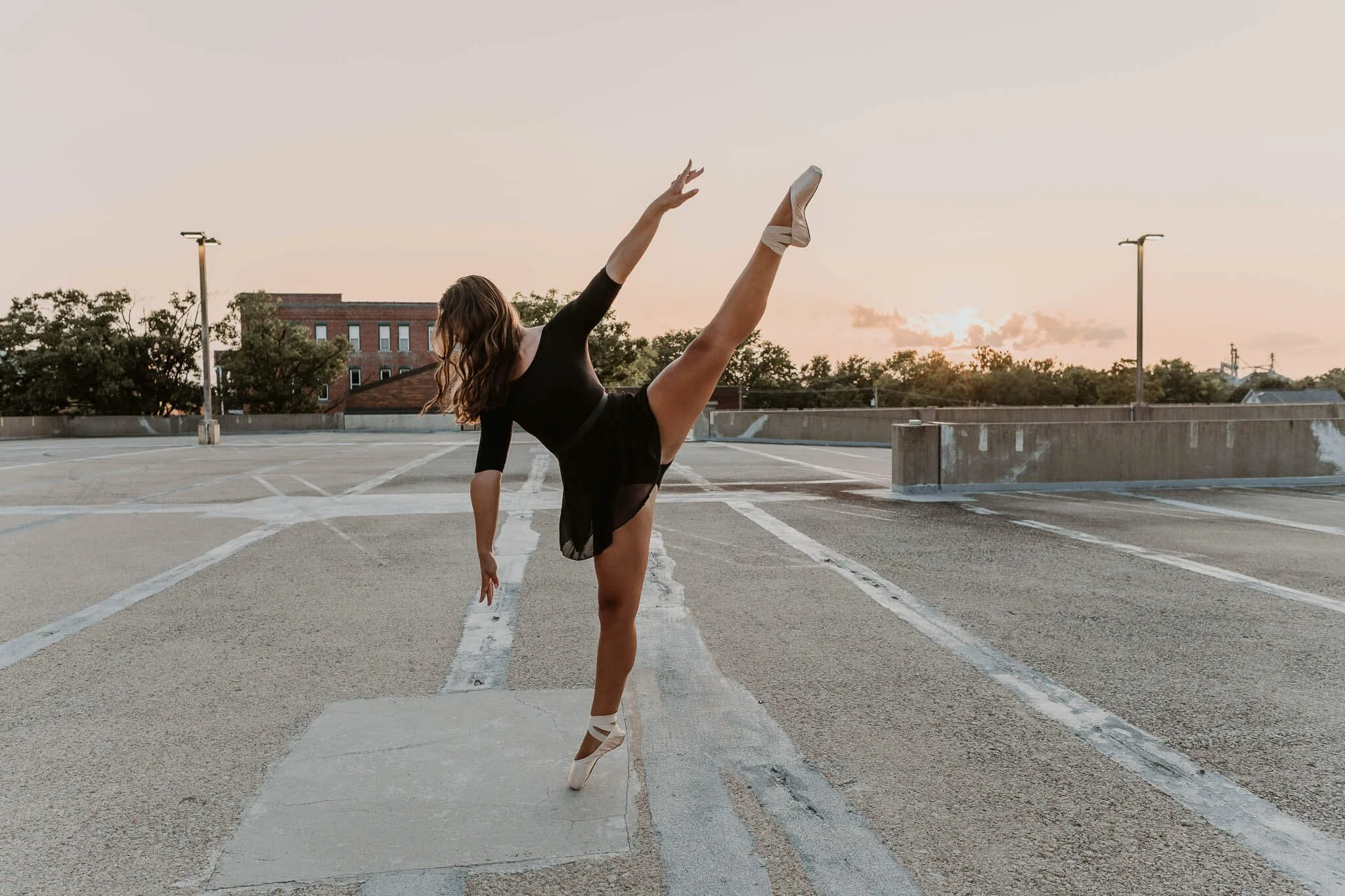 While the sun sets in the background, a talented dancer holds a pose en pointe, hands stretched gracefully above and below her