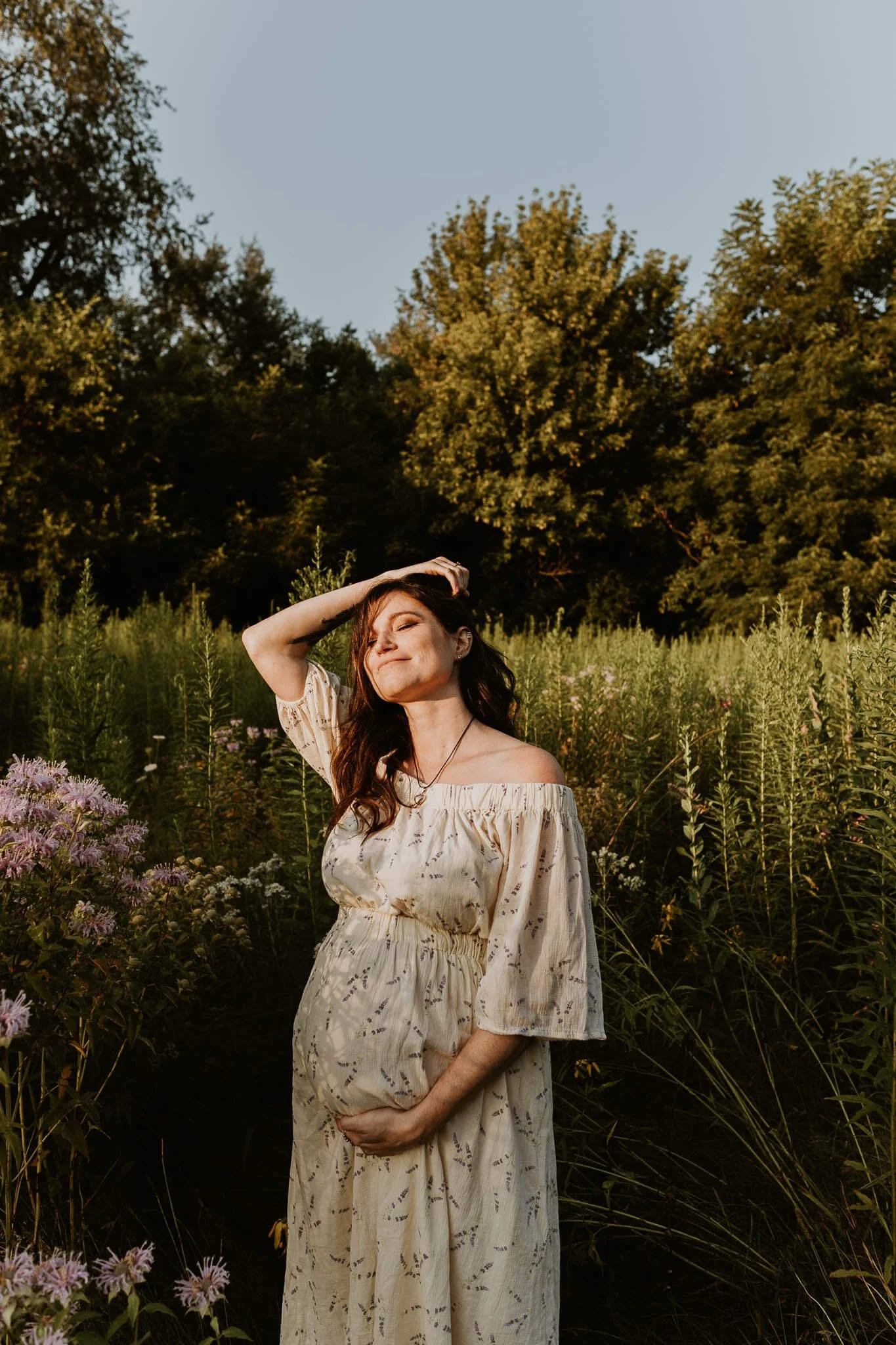 A pregnant mom smiles softly, eyes closed and face turned up towards the sun in a field of wildflowers