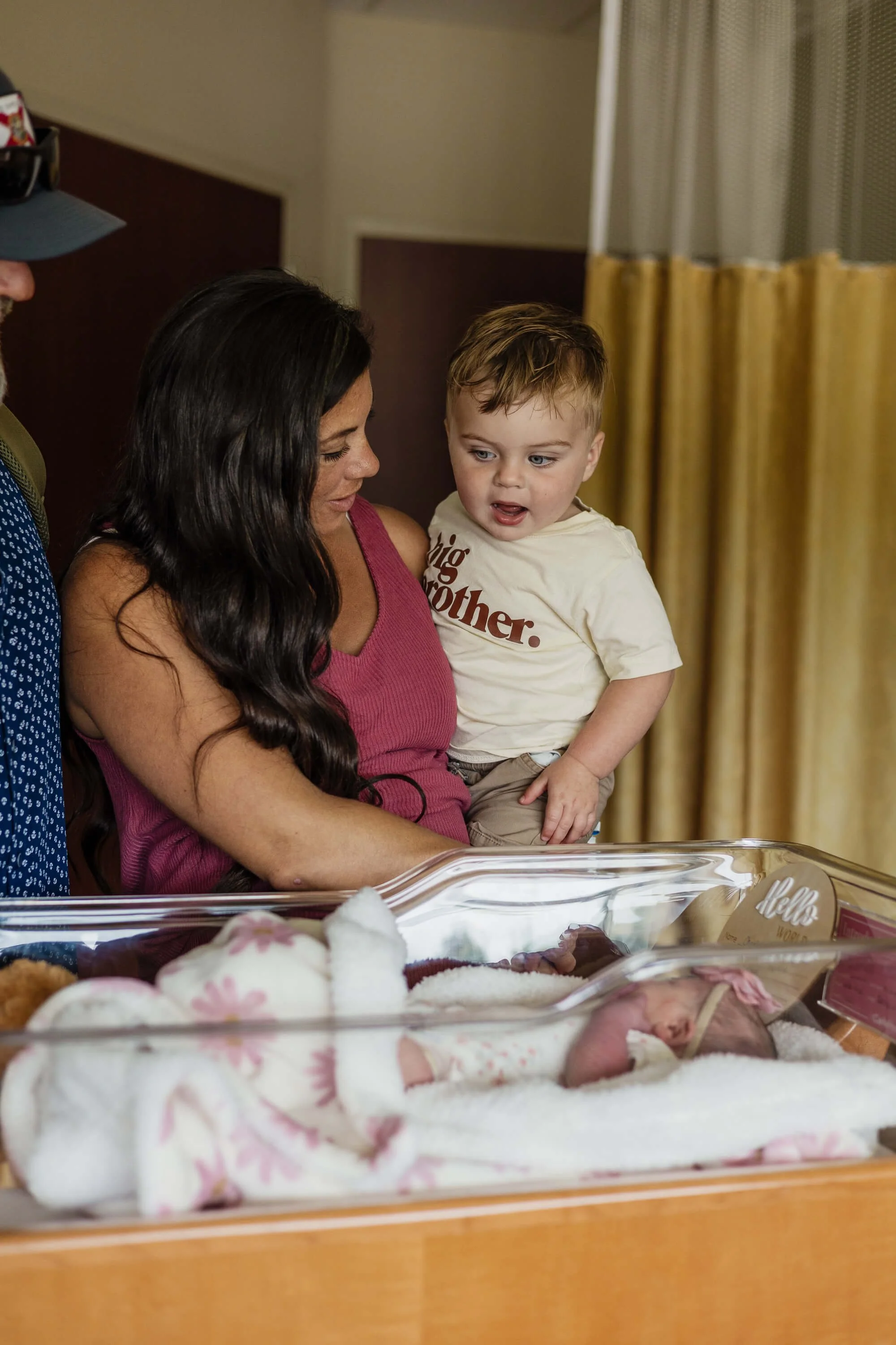 A newly postpartum mom holds her toddler son as they look into his baby sister's hospital cot, his mouth agape in wonder as he stares at his new sister for the first time