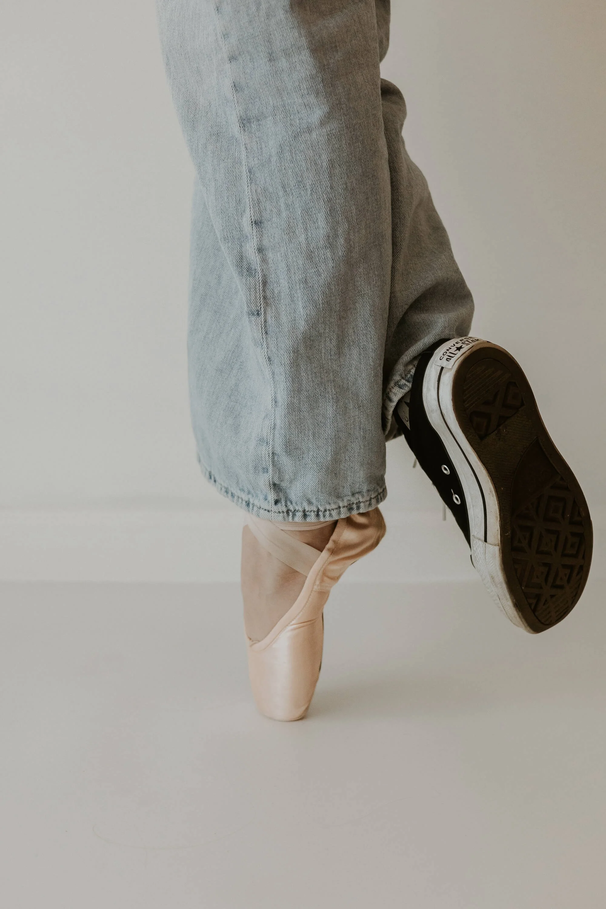 A close up of a ballerina en pointe in her left foot while her right foot dons a classic Converse shoe