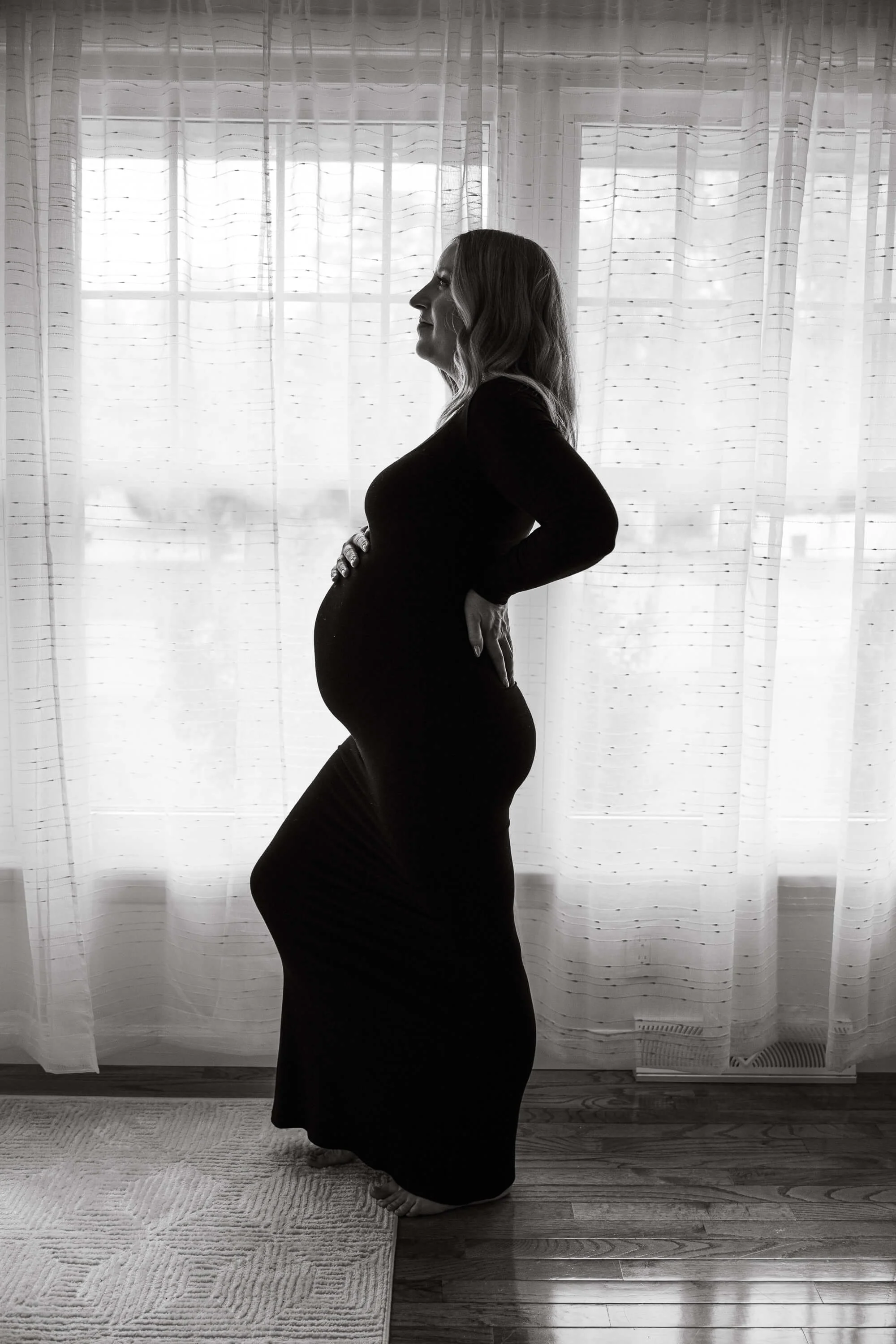 A pregnant mom's sillhouette in front of large windows holding her belly, hand on her back