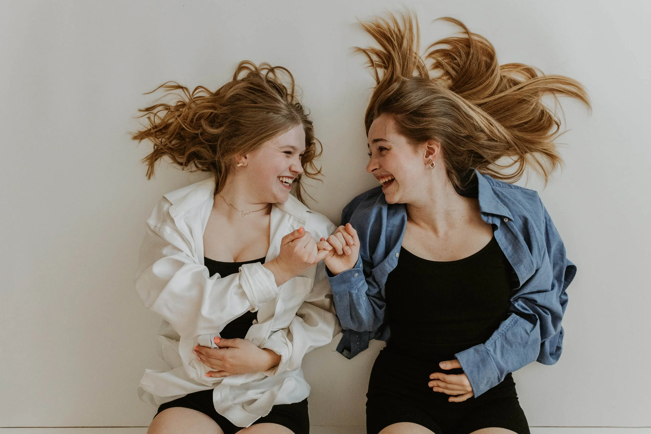 Two teen girls laugh, looking at each other while holding pinkies together. 