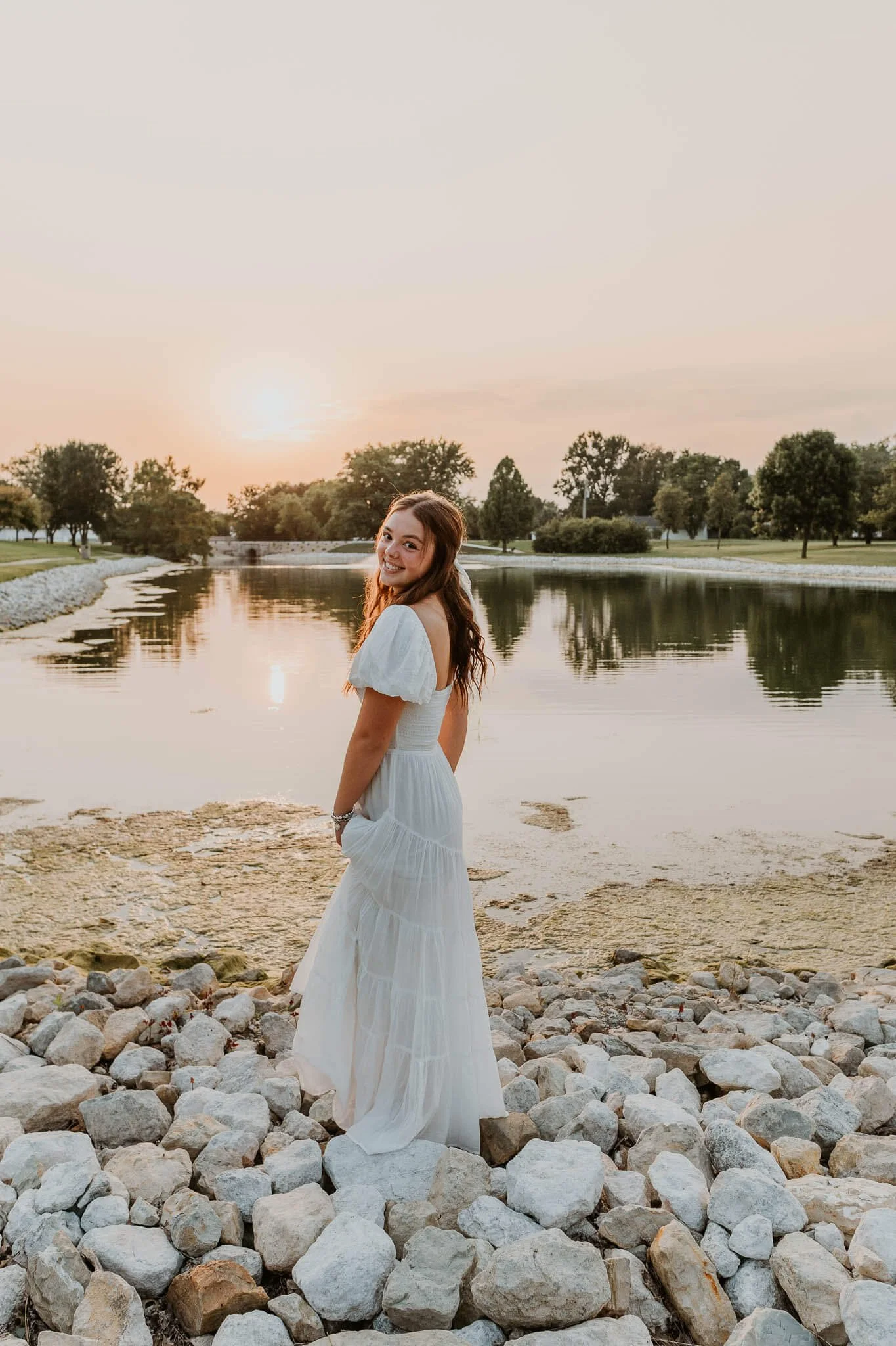 A teen girl wearing a cotton white maxi dress, smiles as she looks over her shoulder atop decorative landscape rocks at a pond.