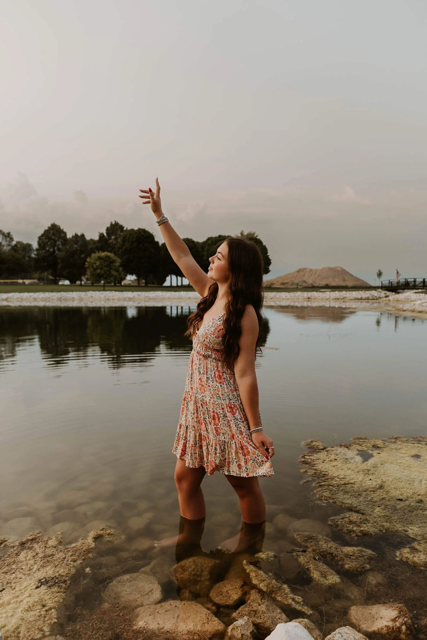 Wearing a floral dress, a senior girl stands peacefully, water lapping at her calves in the pond at Hopedale Park.