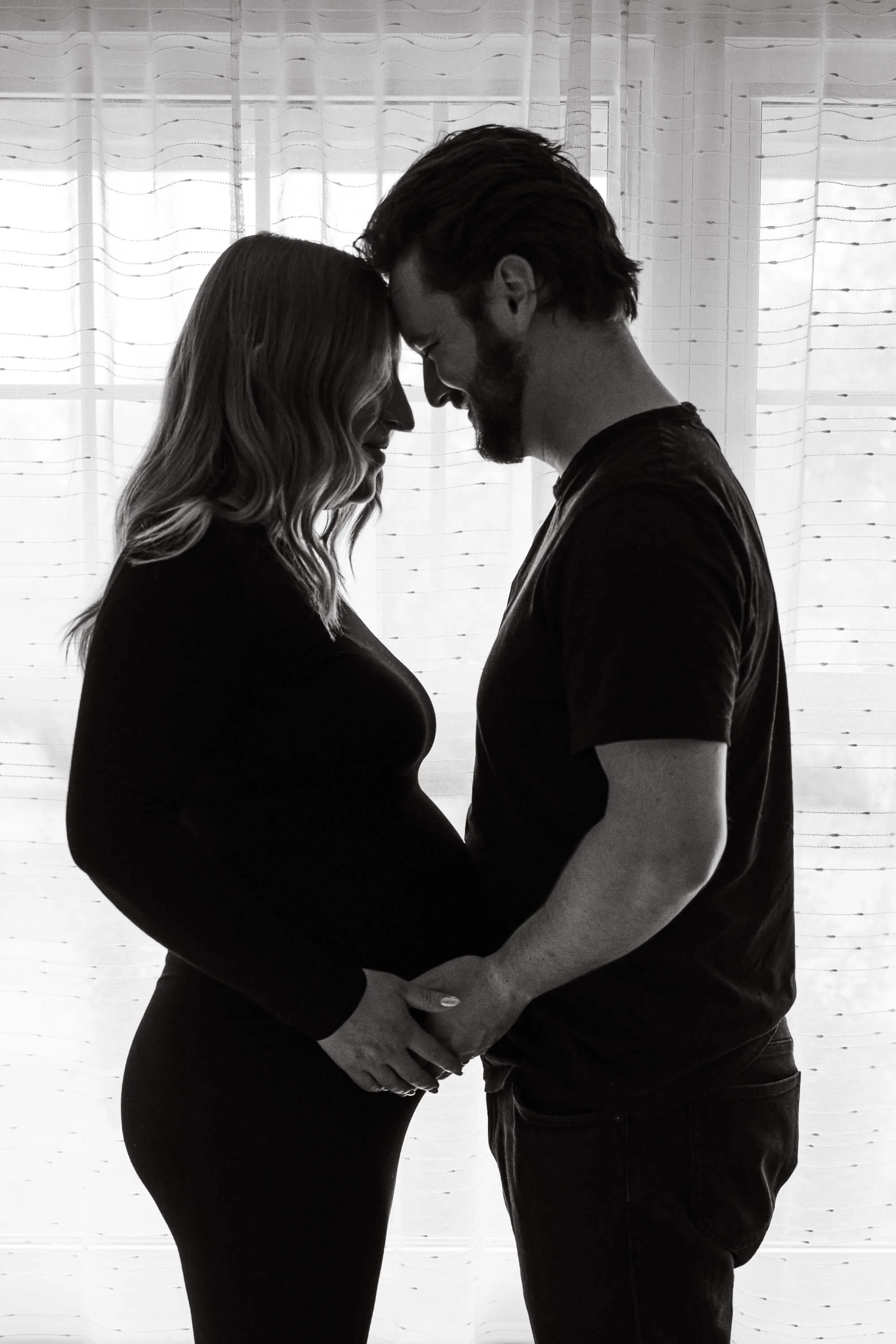 A black and white image of a pregnant woman and her husband holding her belly while facing one another