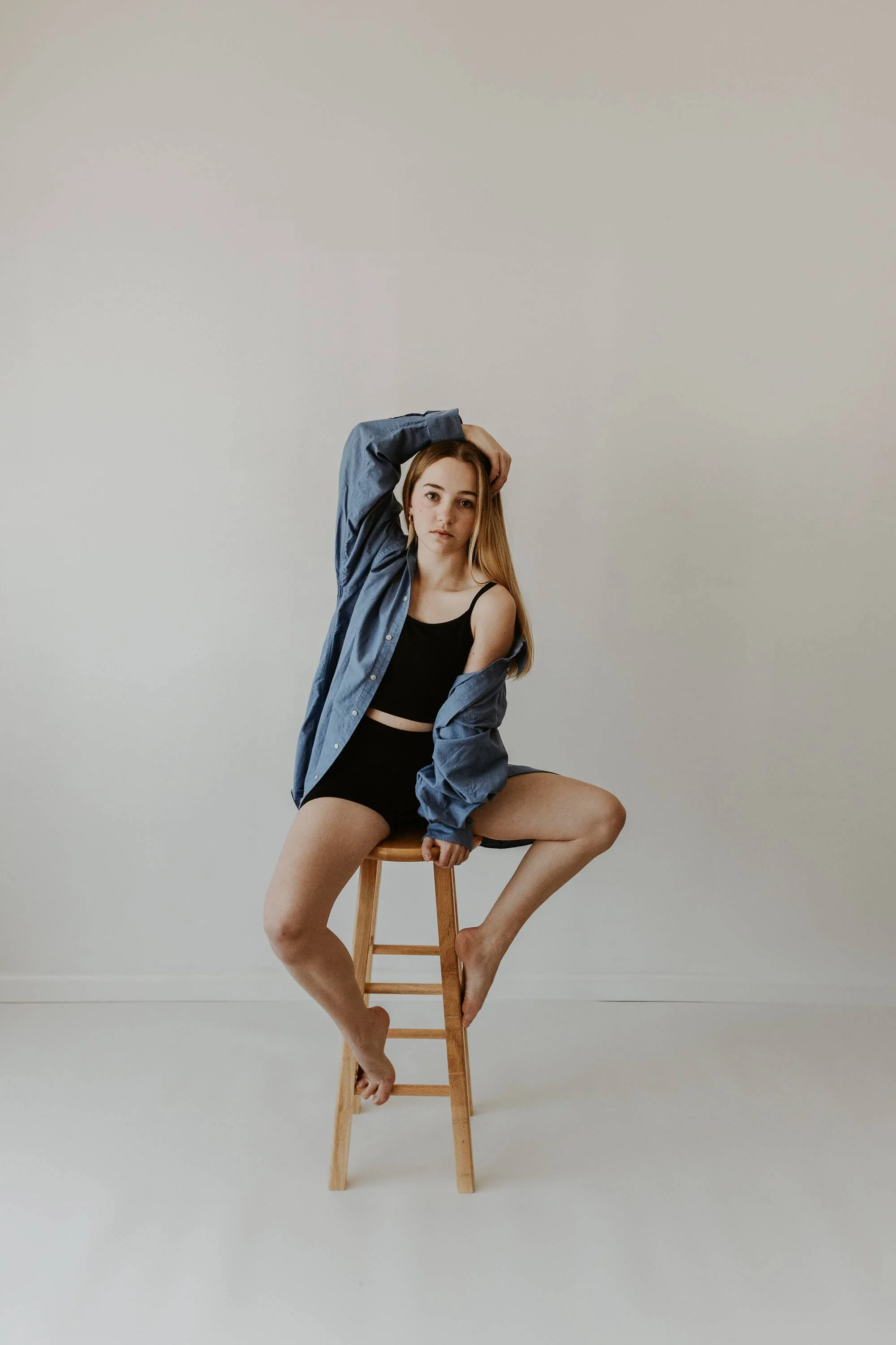 A high schooler holds a pose atop a wooden stool, one hand causally atop her head.