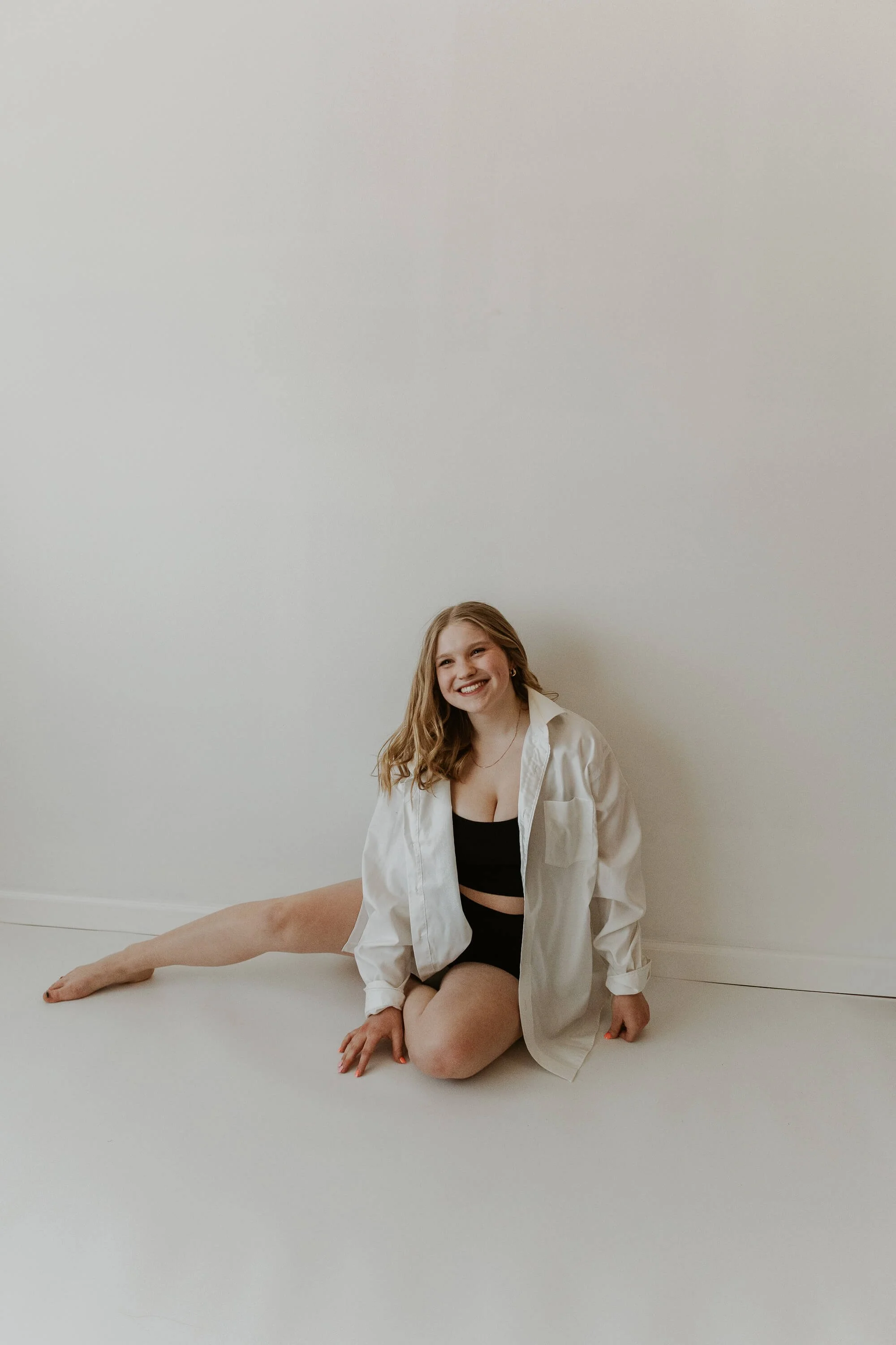 A dancer sits casually against a white wall, softly smiling during her studio session in Bloomington, IL