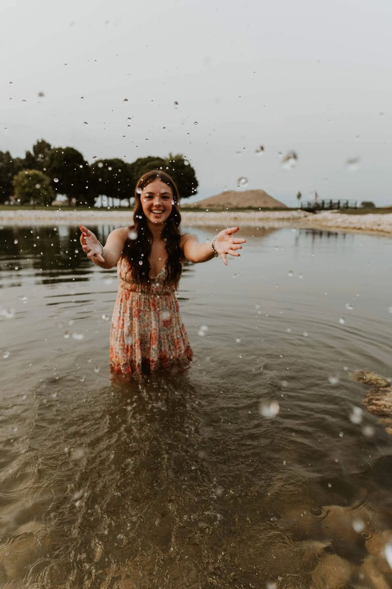 A high school girl playfully splashes water while standing in a pond at a park.