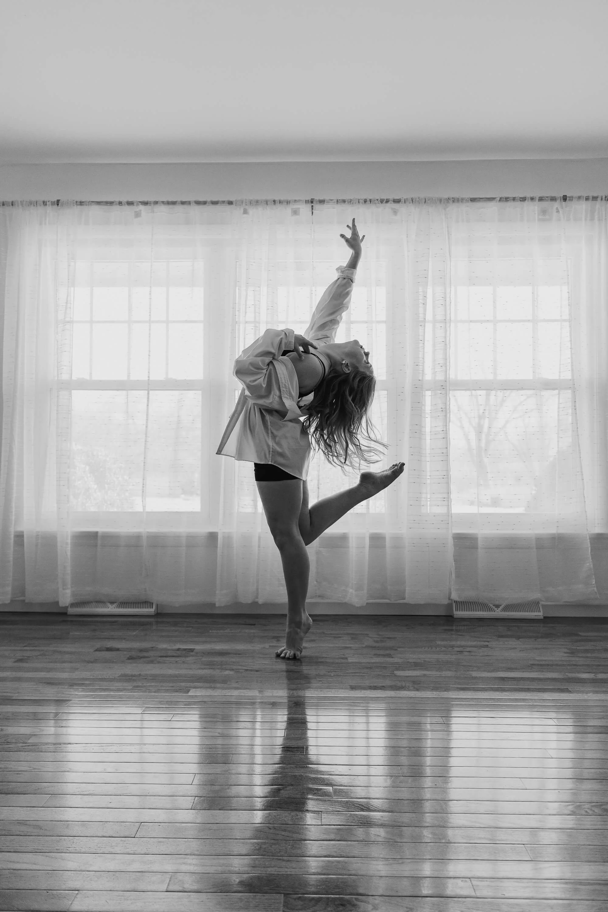 A black and white image of a dancer holding a pose on tip toe in front of a wall of windows.