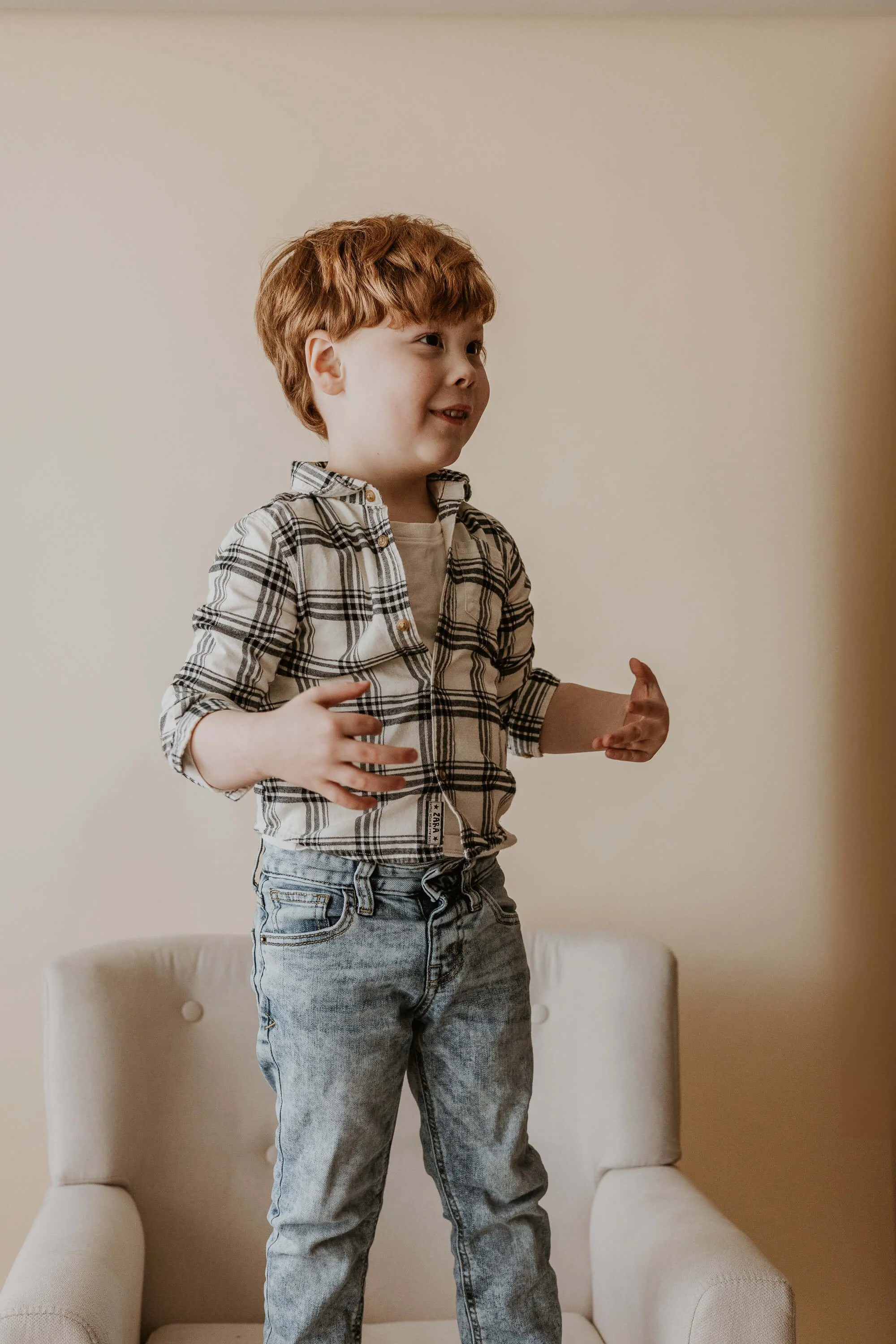 A four year old boy smiles as he stands on a cream chair
