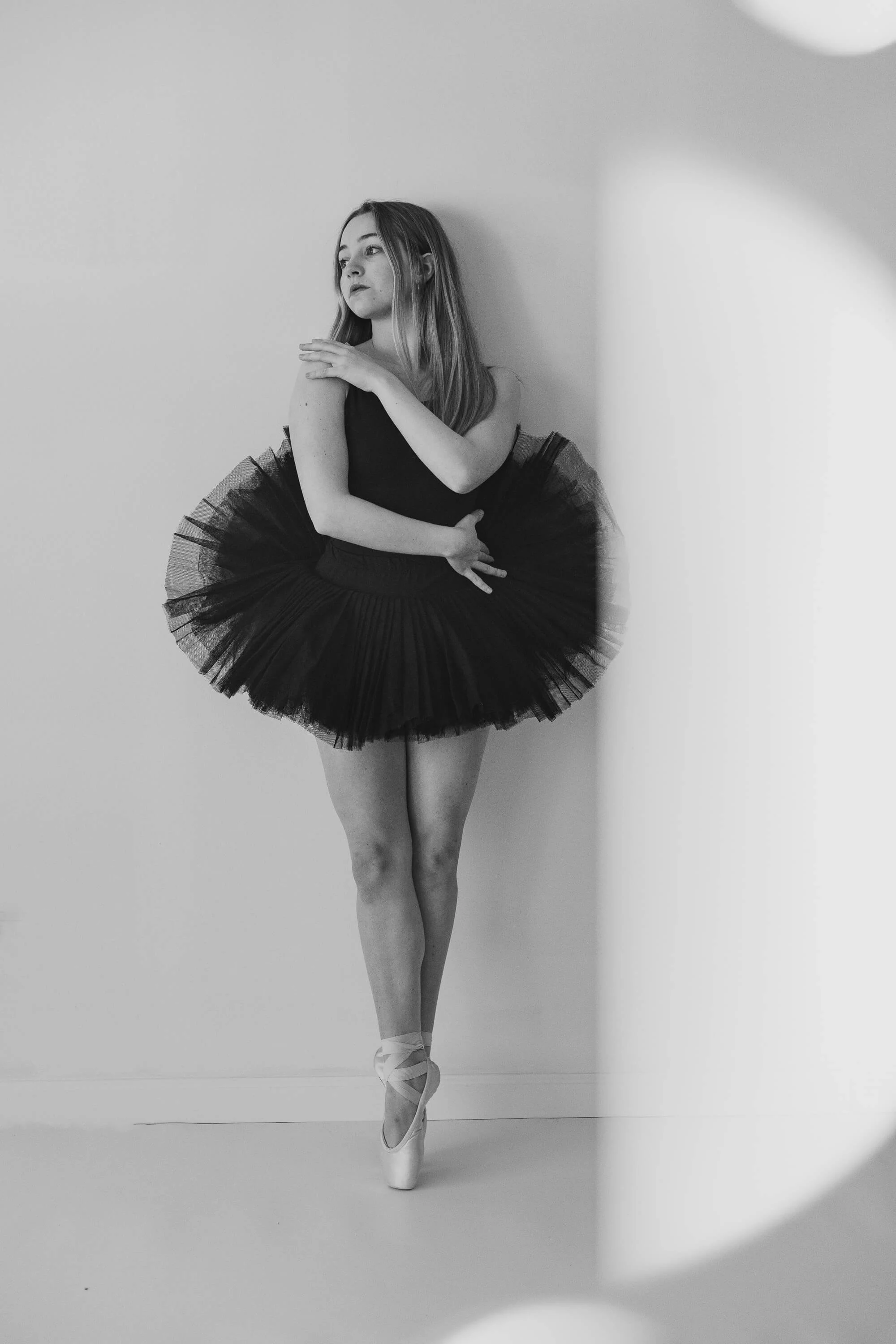 A black and white of a teen girl in ballet shoes, wraps her arms around her mid section and looks off to the side.