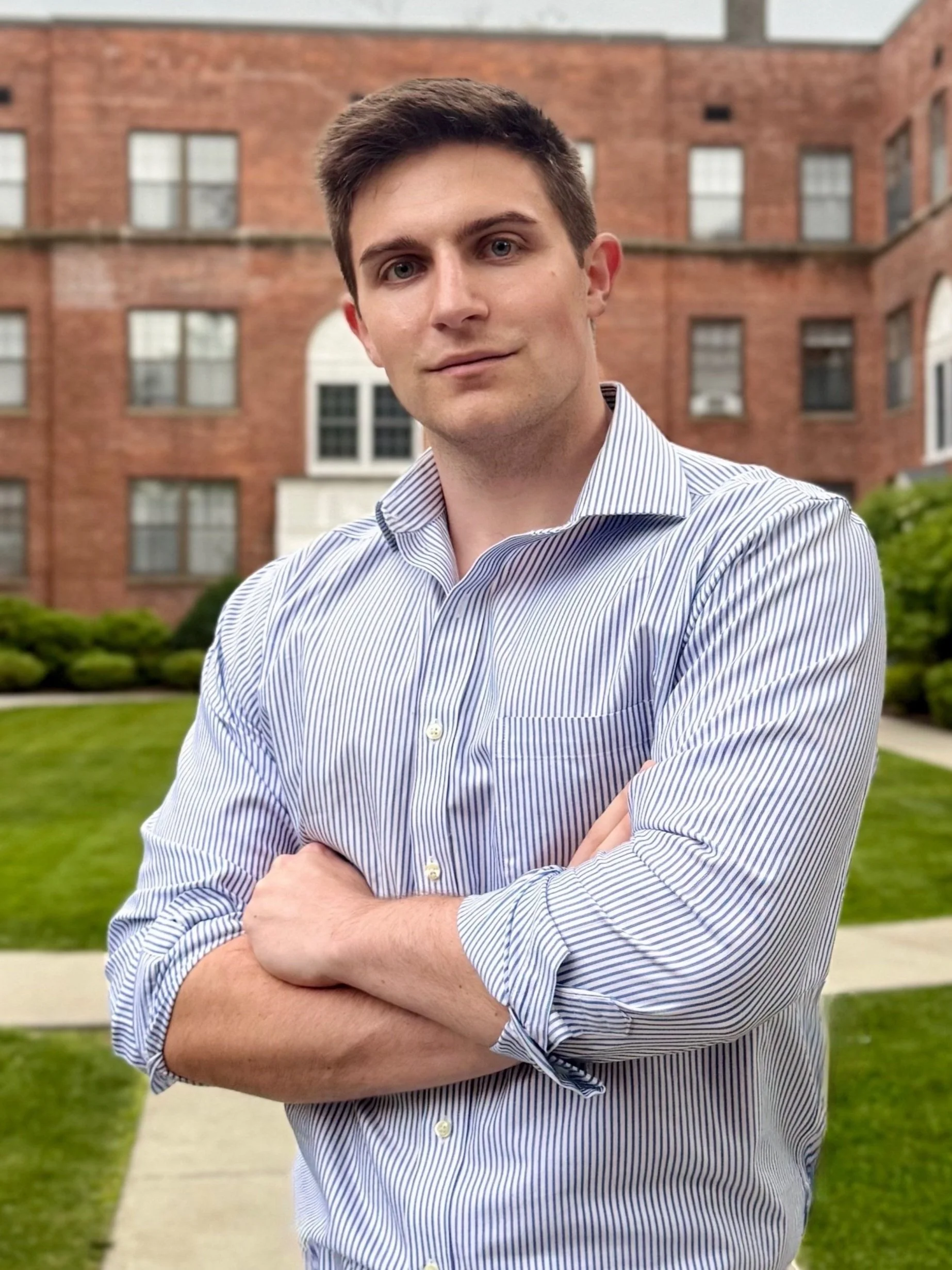 A young man with brown hair and blue eyes, wearing a white and blue striped dress shirt with sleeves rolled up, stands outdoors in front of a brick apartment building and a neatly maintained grassy lawn, with his arms crossed and a confident expression.