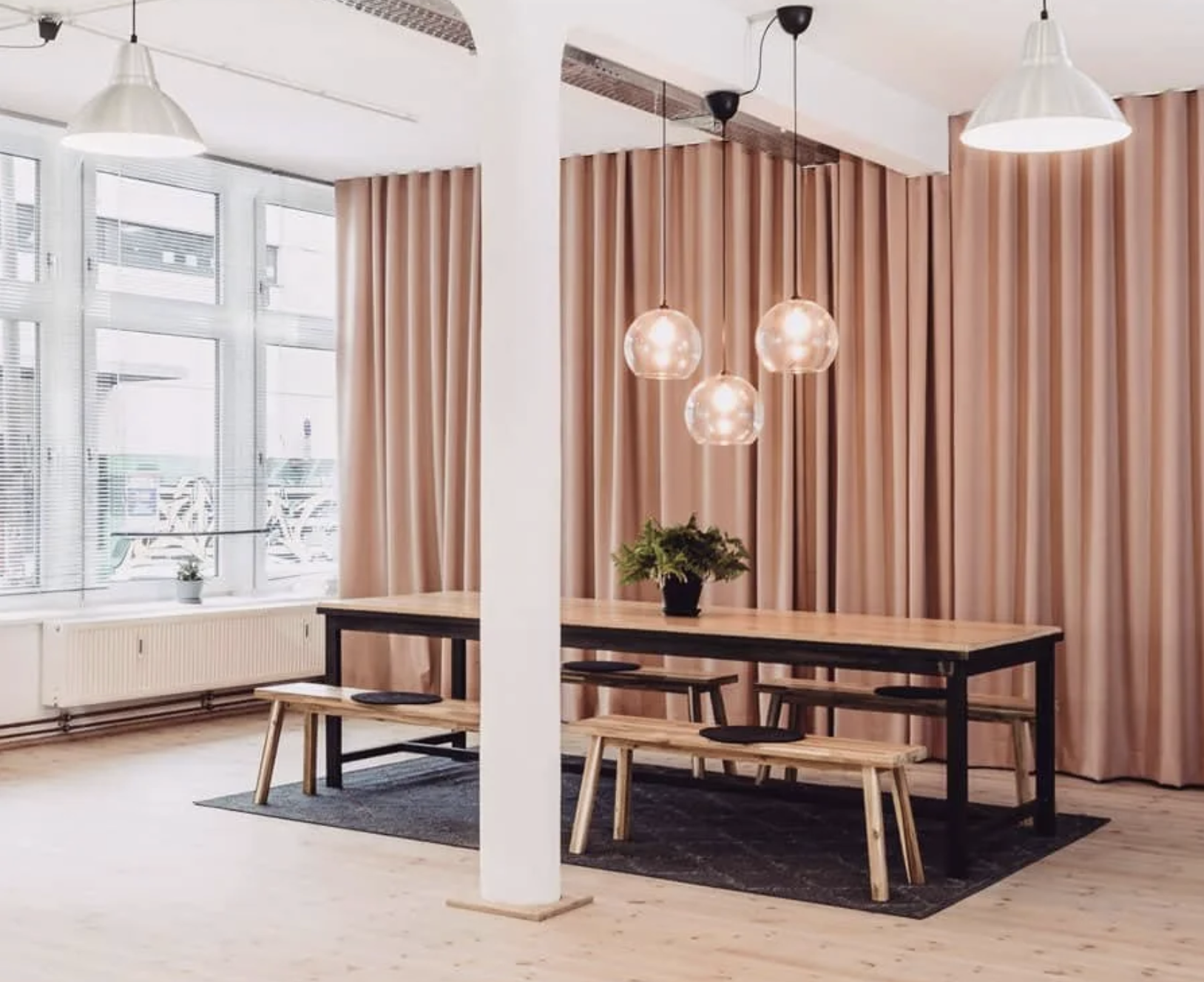 Ein heller Raum mit großen Fenstern, langen pinkfarbenen Vorhängen, einer dunklen Tisch mit zwei Holzbänken, einer kleinen Pflanze auf dem Tisch und vier hängenden Glühbirnen. Der Raum hat Holzböden und eine weiße Säule.