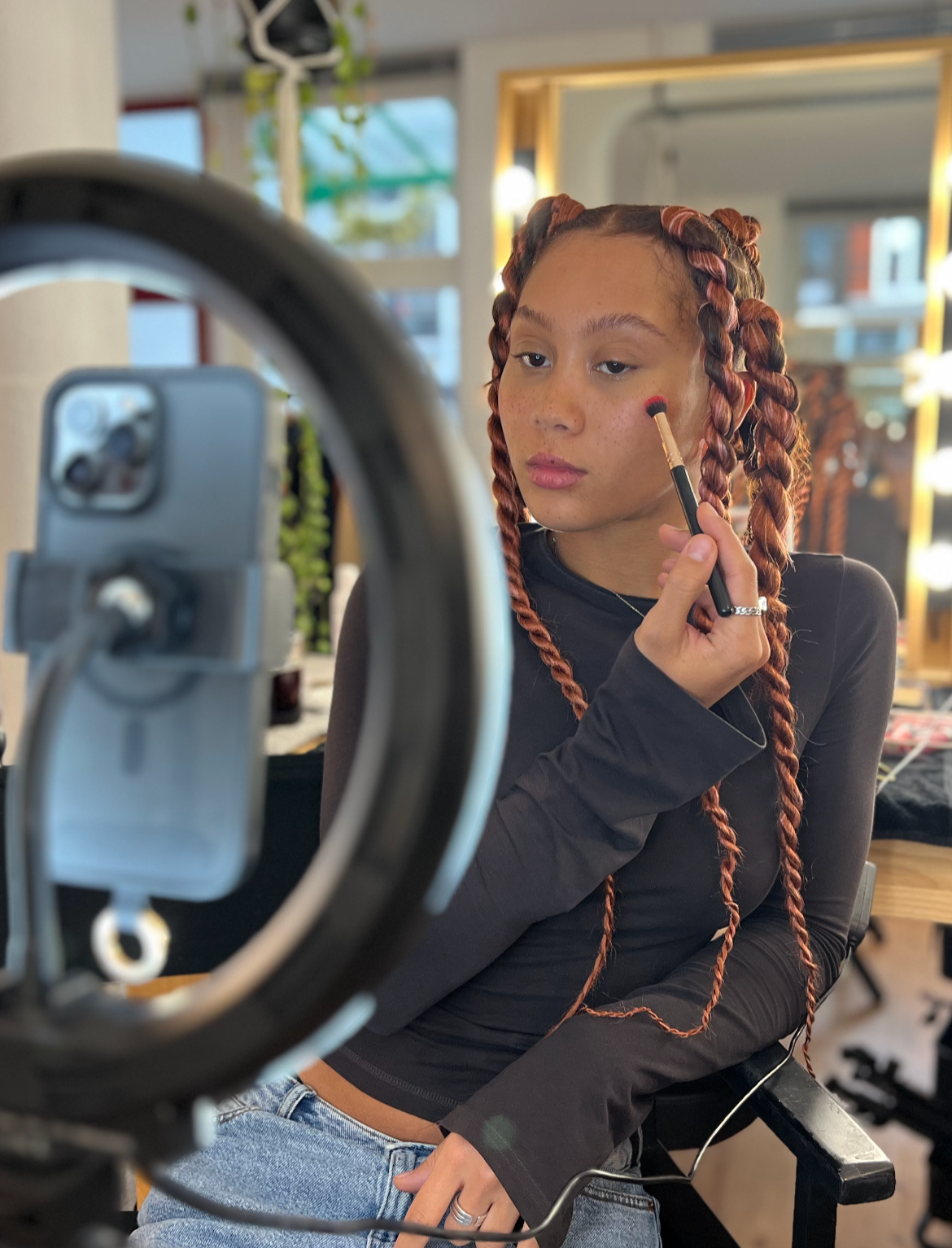 Junge Frau mit geflochtenen Haaren, die Make-up aufträgt, vor einem Spiegel mit Beleuchtung in einem Beauty-Studio.