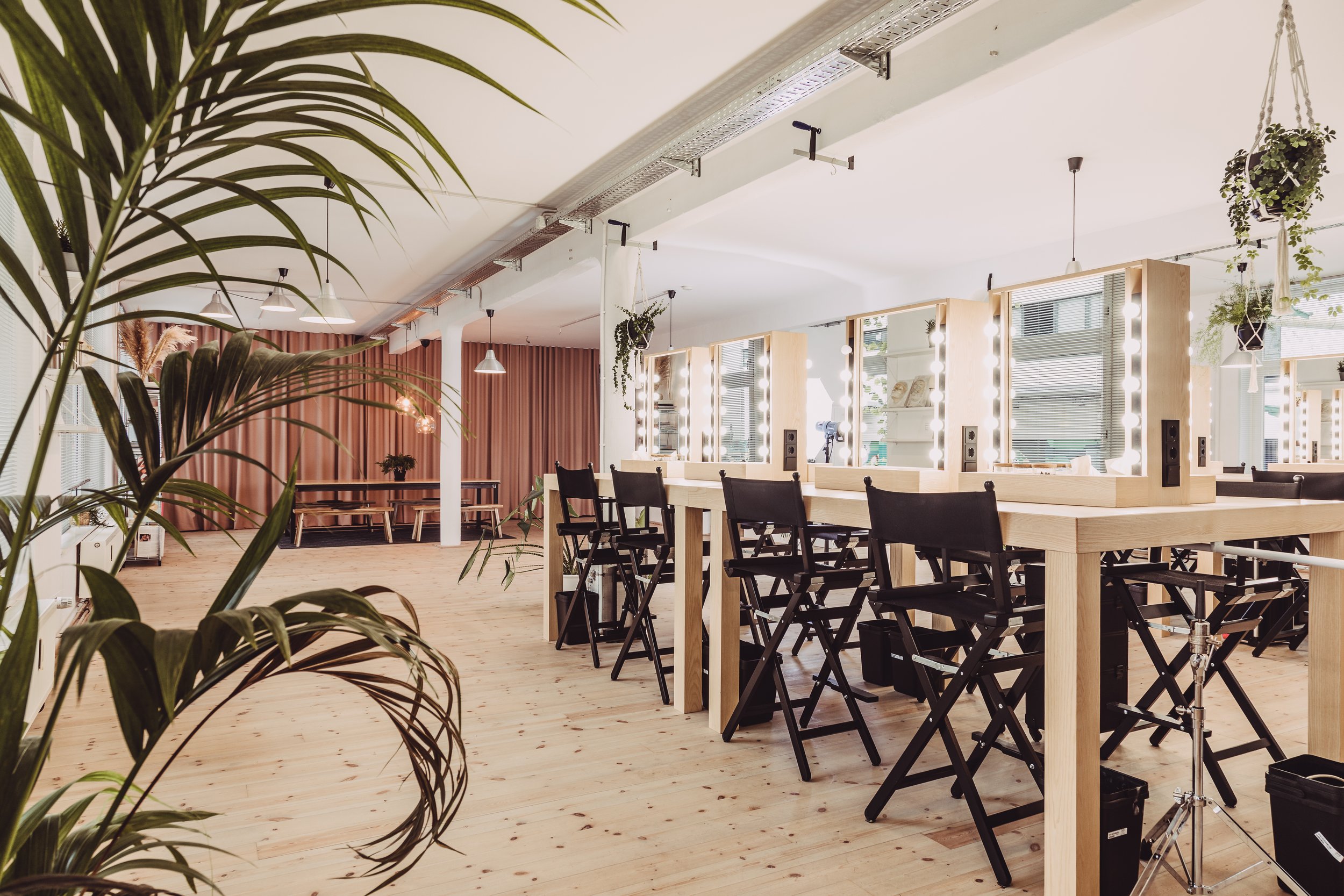 Ein schön eingerichteter Make-up-Raum mit beleuchteten Spiegeln, schwarzen Stühlen und einigen Pflanzen, mit einem natürlichen Holzdesign und einem hellen, freundlichen Ambiente.