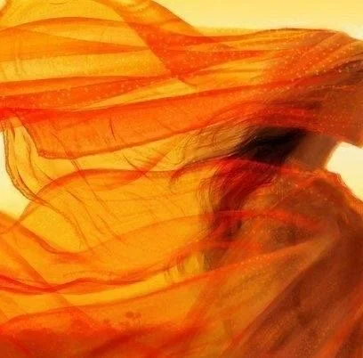 Flowing orange sheer fabric swirling around a woman in soft golden light, symbolising the rising of buried emotion, the movement of the Holy Spirit, and the prophetic fire that precedes resurrection.