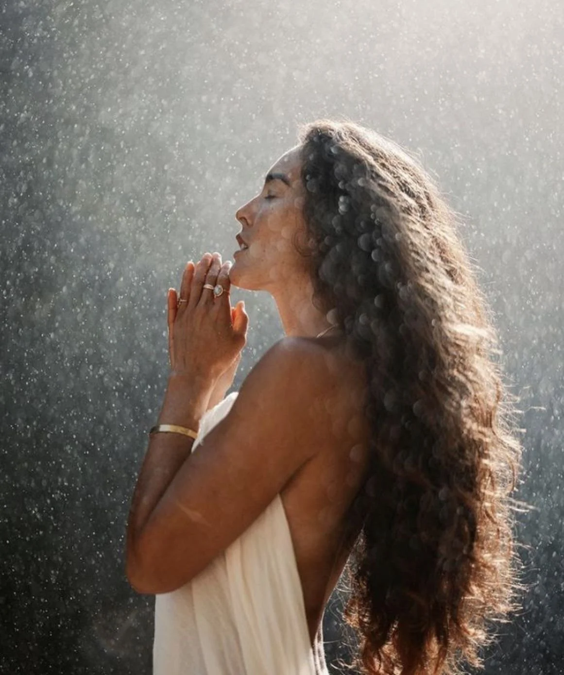 A woman standing in soft rainlight with her eyes closed and hands in prayer, illuminated by the Holy Spirit’s presence, symbolising the physical and spiritual power of divine encounter.