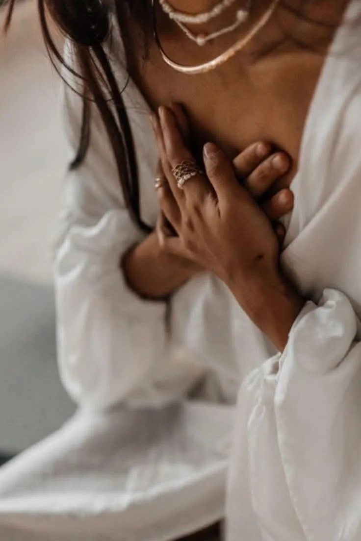 Close-up of a woman in a white robe with hands placed over her heart, adorned in delicate rings and layered necklaces, symbolising reflection, devotion, and heartfelt connection.