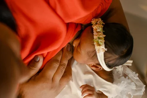 A mother and baby with dark skin breastfeed. The baby wears a headband with flowers.