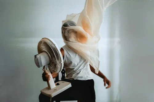 A person stood holding a fan up to their face, with a  sheer scarf blowing around them