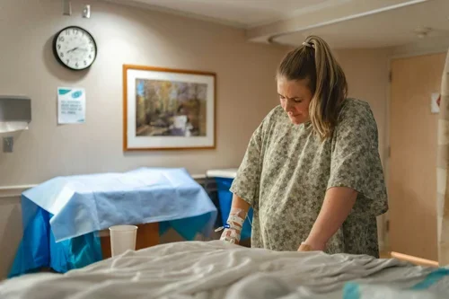 Pregnant person, who is white with long hair in a pony tail and wearing a hospital gown, stands in a room on a maternity unit.