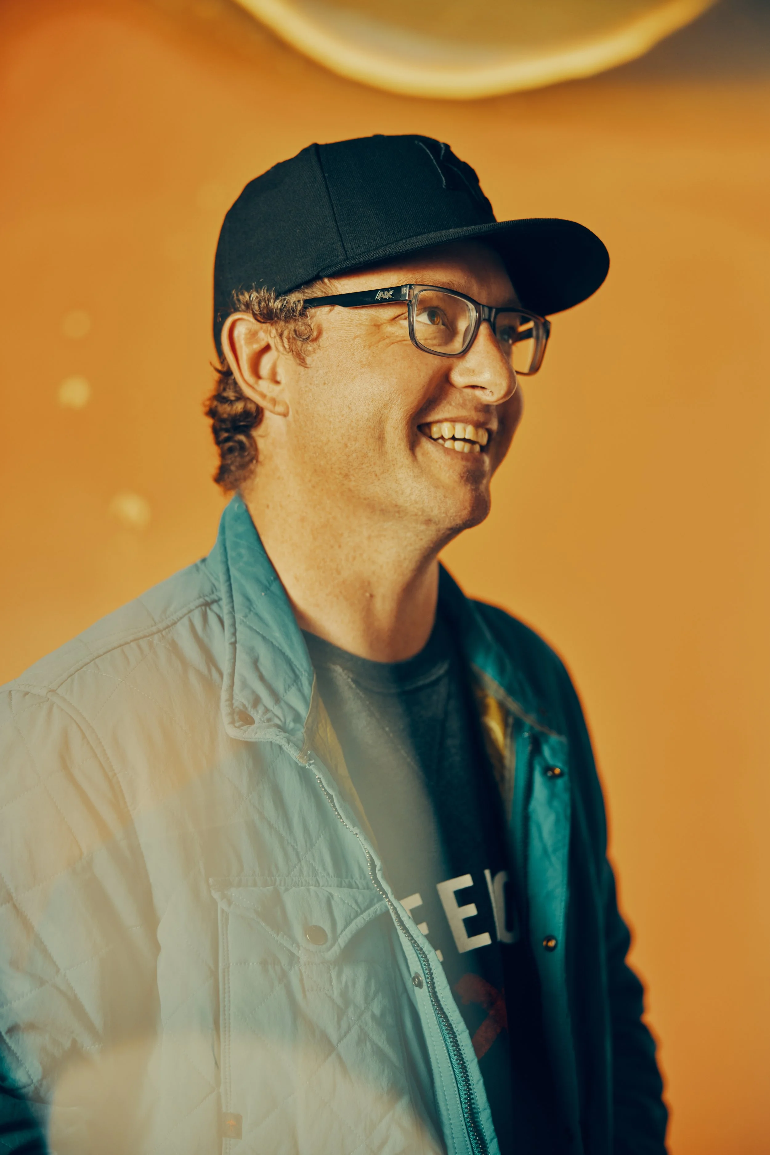 A man smiling, wearing a black cap, glasses, a blue jacket, and a black t-shirt with white lettering on an orange background.
