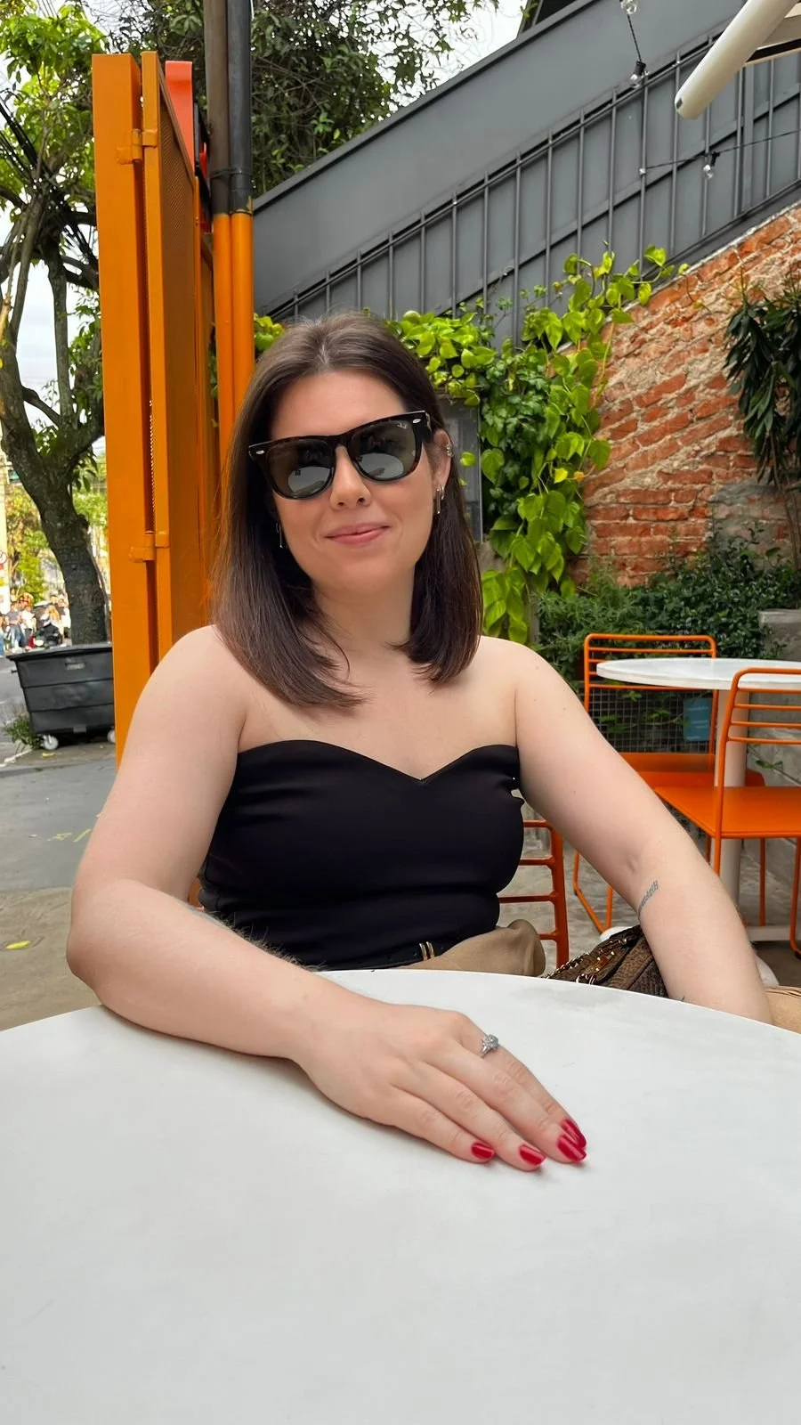 Mulher de cabelo castanho, usando óculos escuros, camiseta preta e anel, sentada ao ar livre em um restaurante, com fundo de plantas verdes, parede de tijolos e móveis laranja.