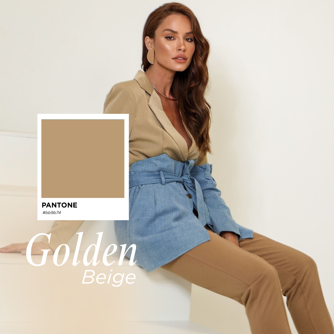 Mulher com cabelo castanho e olhos castanhos usando blusa bege, saia jeans azul e calças bege, sentada contra parede branca, com jóias visíveis e um fundo branco minimalista.