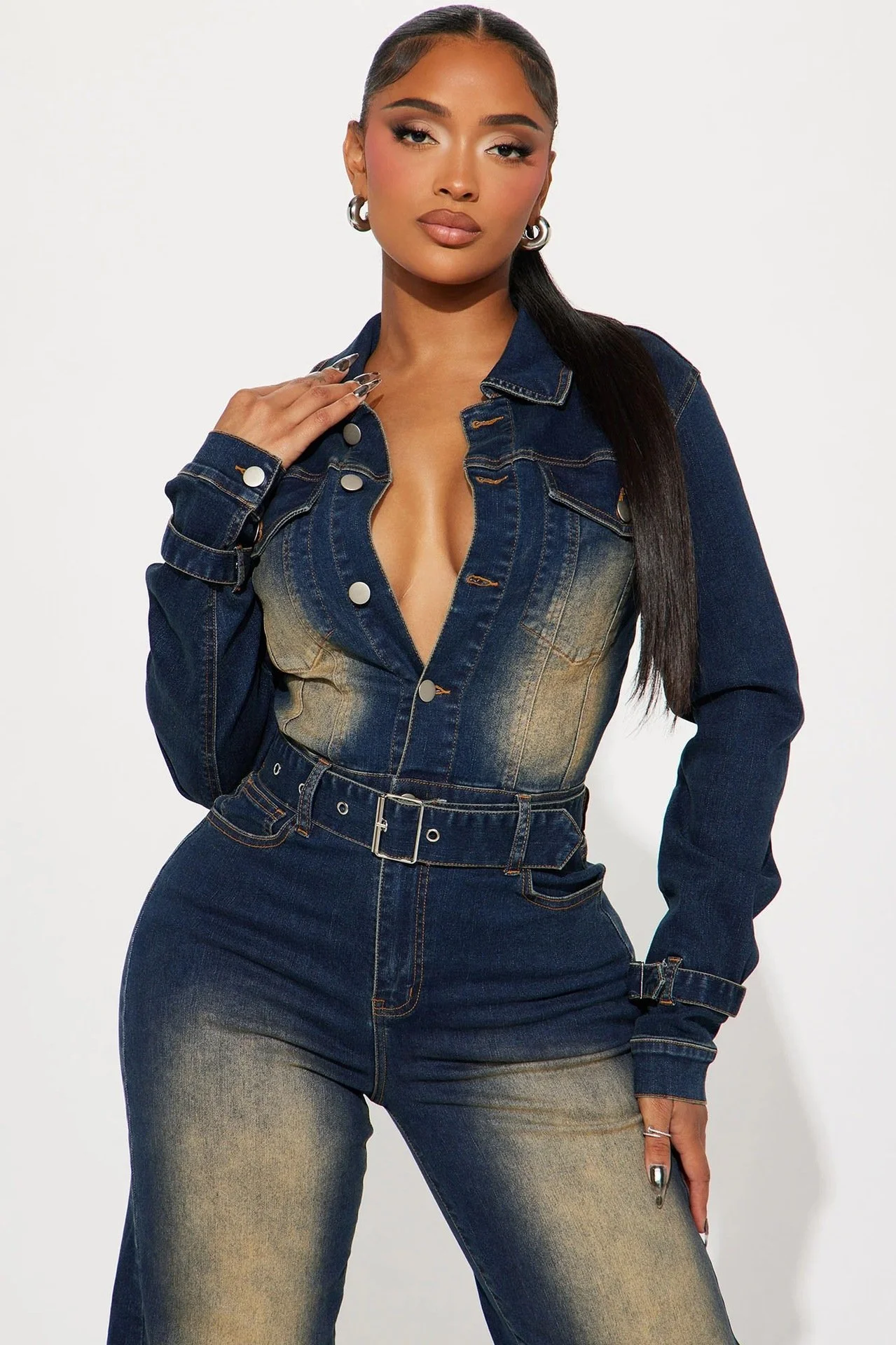 A woman with long dark hair, wearing hoop earrings, a denim jacket, and matching high-waisted jeans, posing against a white background.