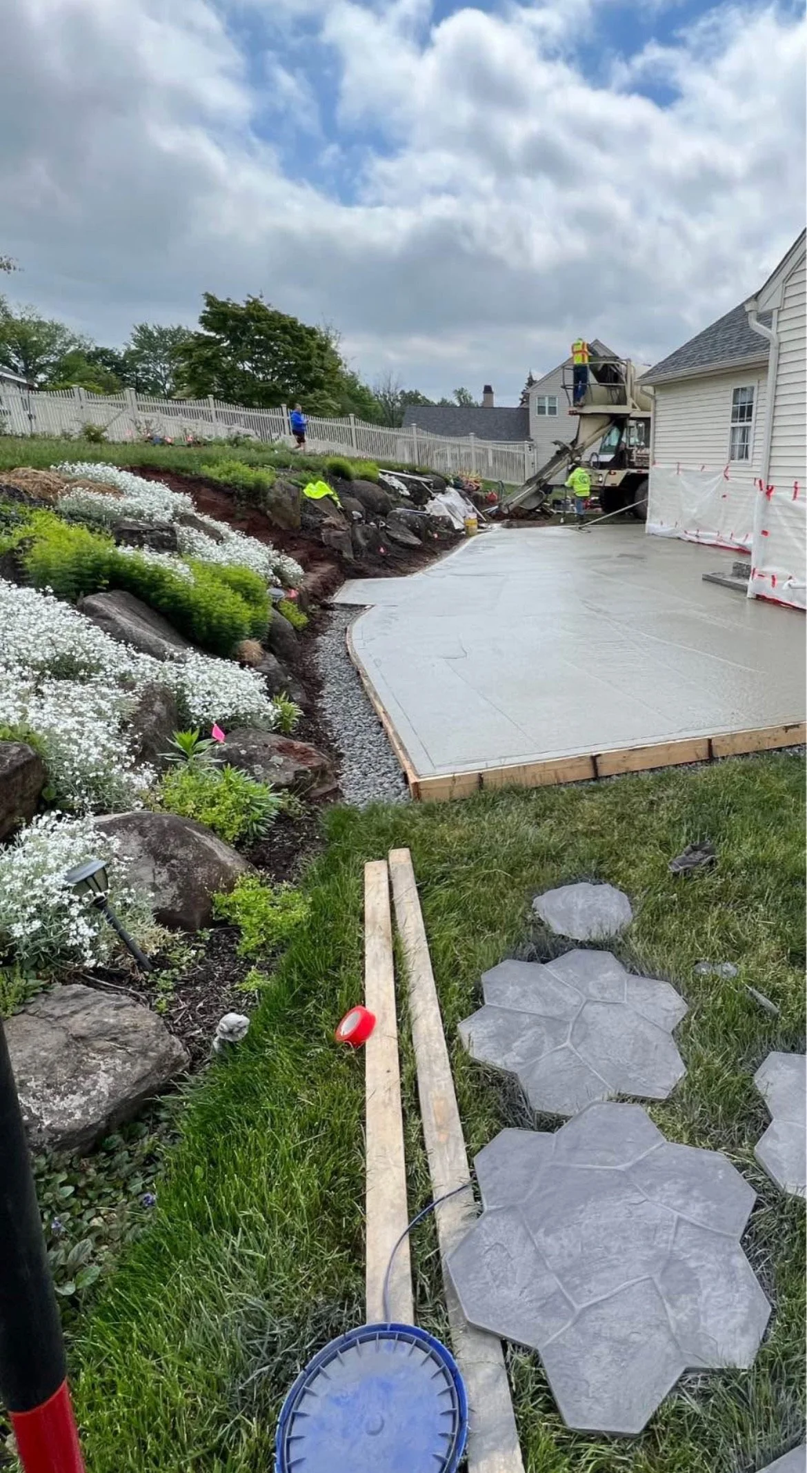 Workers pouring and smoothing concrete for a backyard patio, with landscaping and flowering plants along a hillside, and stone pathway pieces nearby in Hampton new hampshire