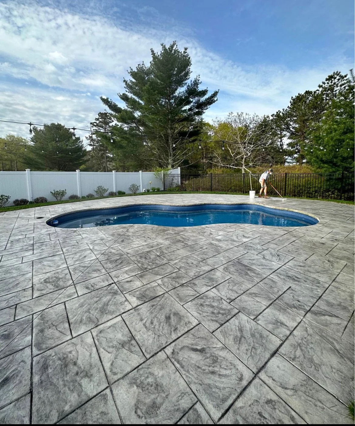 A backyard with a swimming pool, surrounded by a patio with stamped concrete, a white privacy fence, and trees in the background, with a person cleaning the pool area in Groton Massachusetts