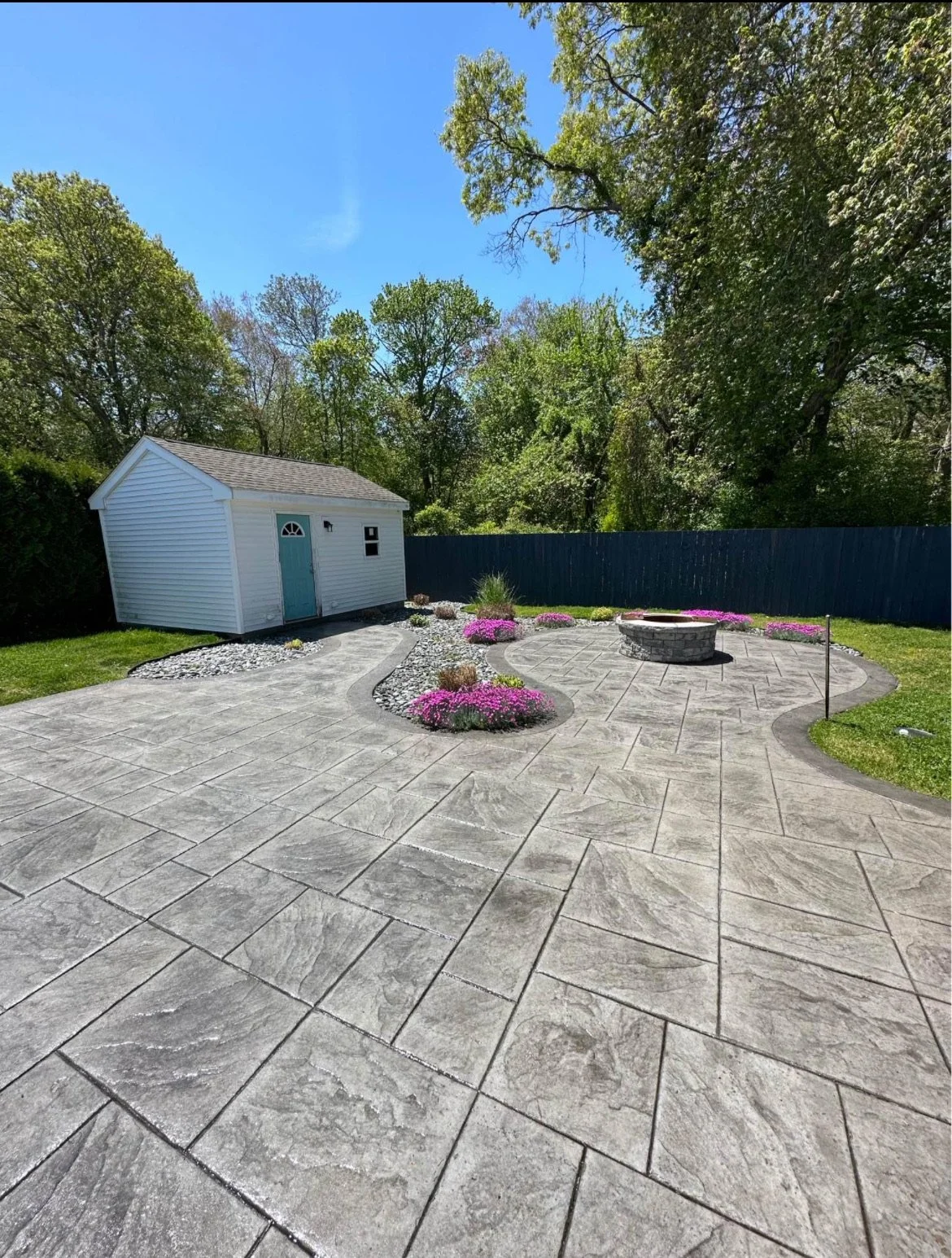 Backyard with a stamped concrete patio, a stone fire pit, a white garden shed, landscaped flower beds with pink flowers, gravel pathways, a blue fence, and trees under a clear blue sky in Northwoods New Hampshire.