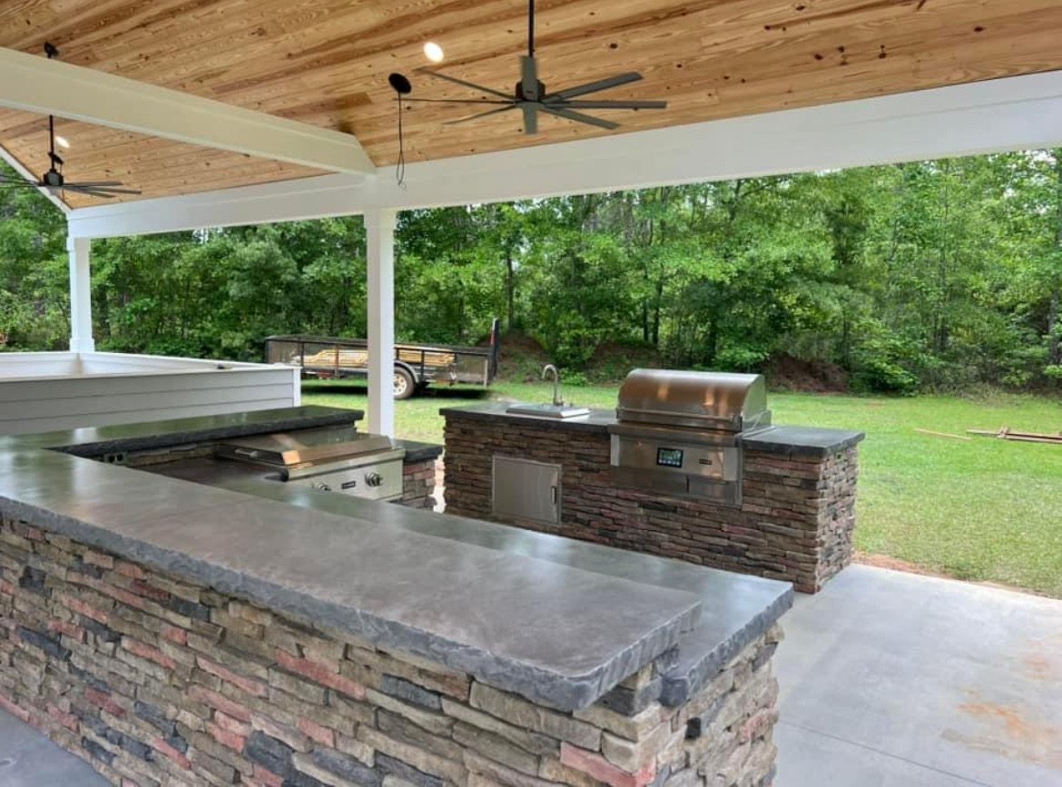 Open outdoor kitchen with decorative concrete stone countertops, stainless steel grill, sink, and ceiling fans, overlooking a grassy backyard with trees in Meredith New Hampshire.