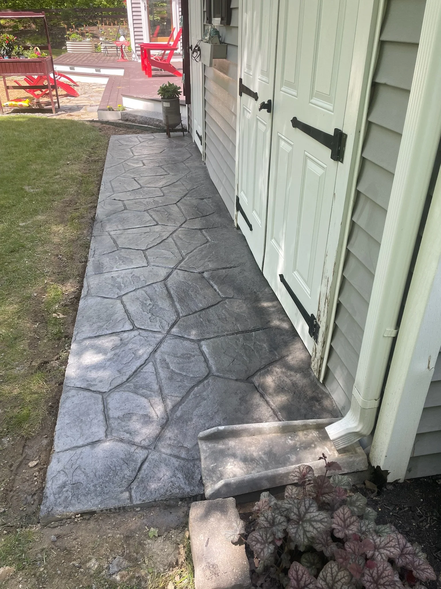 Concrete sidewalk beside house with white shutters, garden with plants and outdoor furniture in backyard in Laconia New Hampshire.