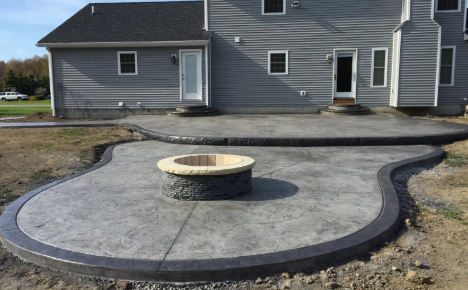 Newly constructed backyard stamped concrete patio with borders and a fire pit, adjacent steps leading to house doors, and surrounding landscaping in Concord New Hampshire.